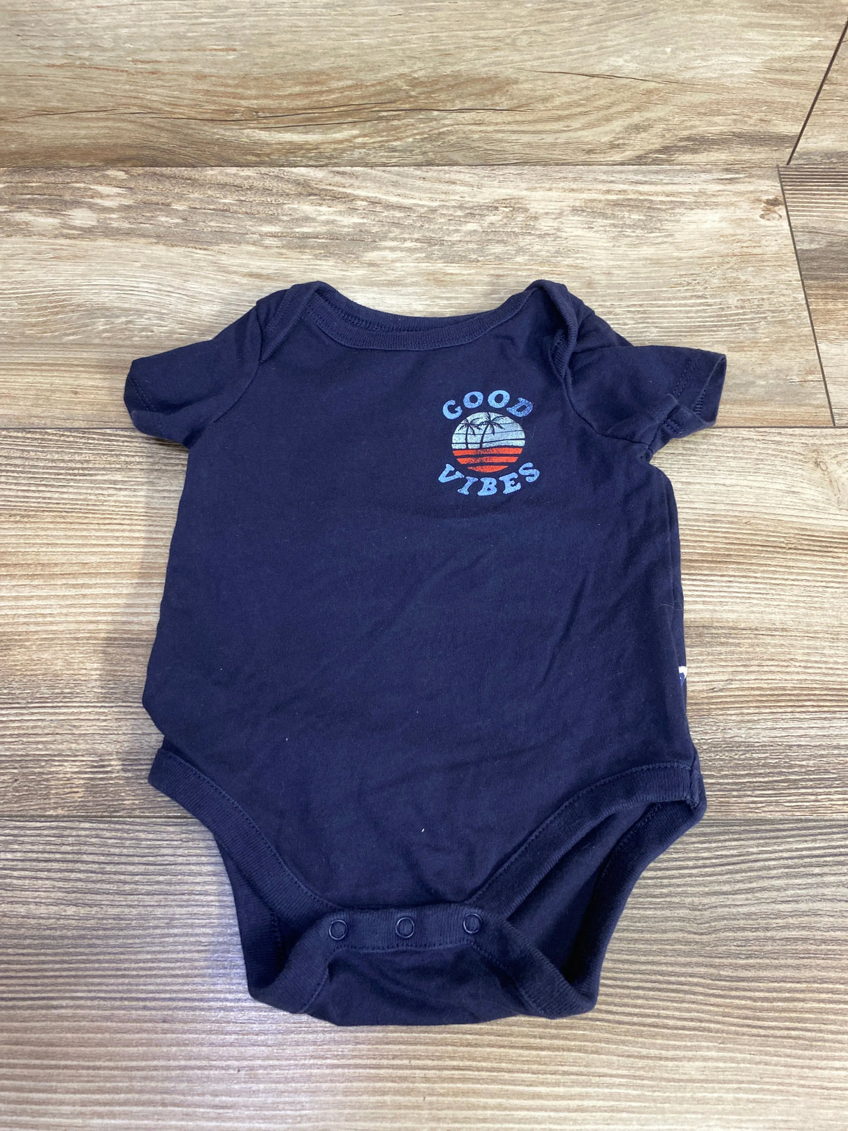 Navy blue baby onesie with "Good Vibes" and palm tree sunset graphic on wooden floor