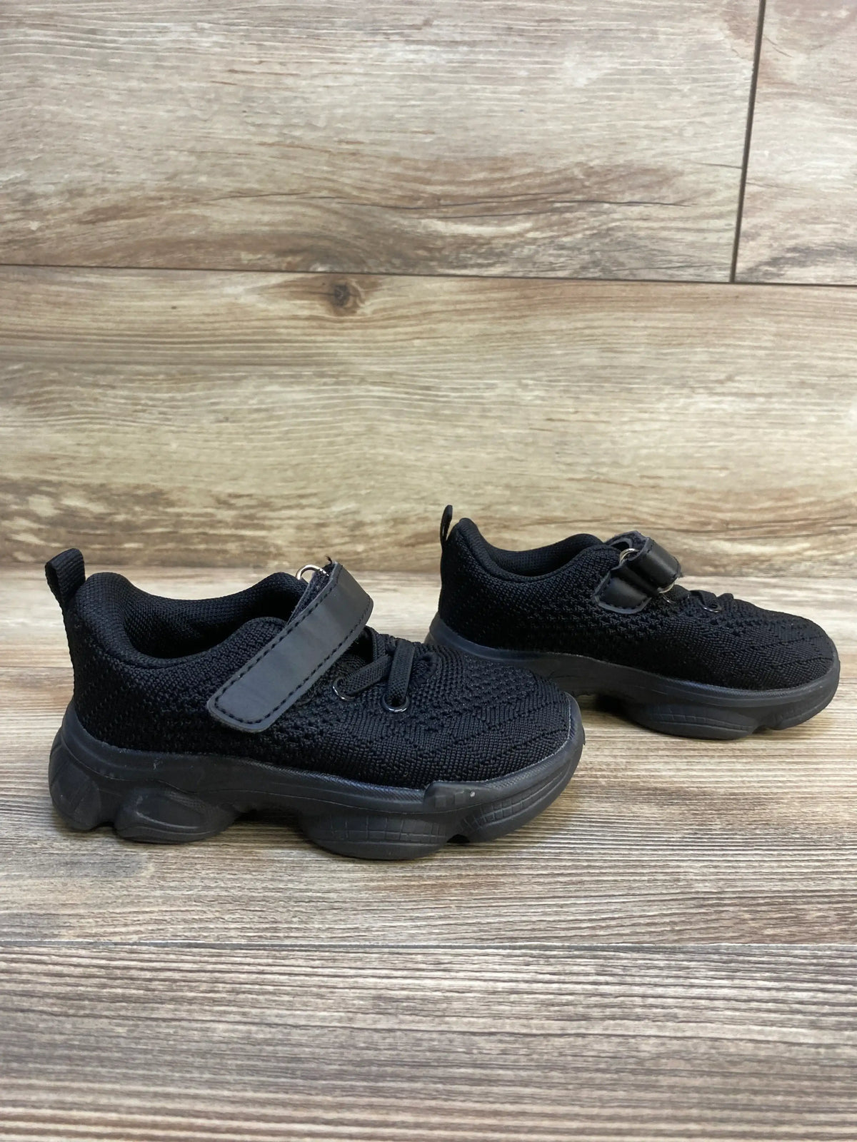 Side view of pair of black Anlarach mesh toddler shoes showcasing Velcro strap, knit upper and chunky rubber sole on wood floor