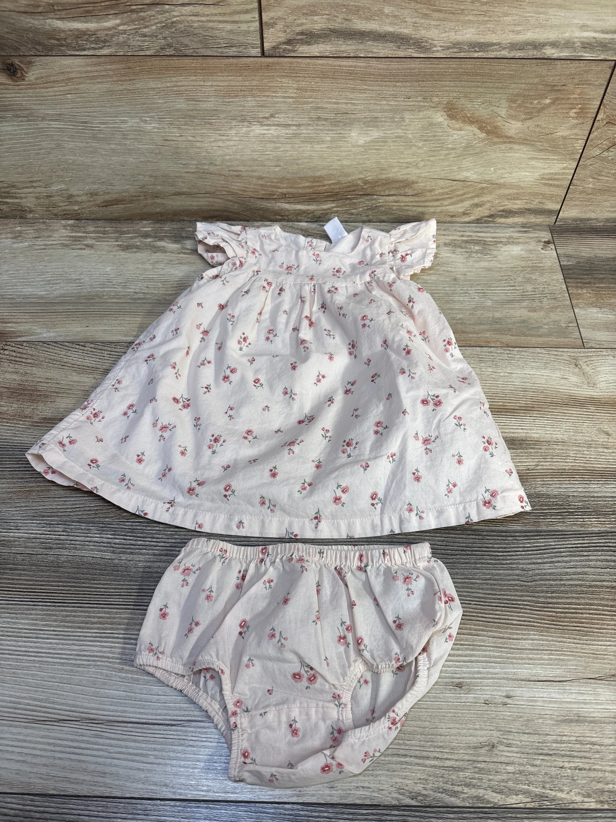 Flat lay of pink floral baby dress and matching bloomers showing ruffle sleeves and gathered skirt on wood floor background
