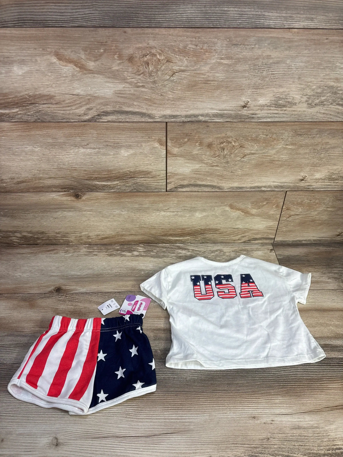 Flat lay of Minnie Mouse 2-piece set: white tee with USA print on back and matching flag shorts, size 18m on wood background