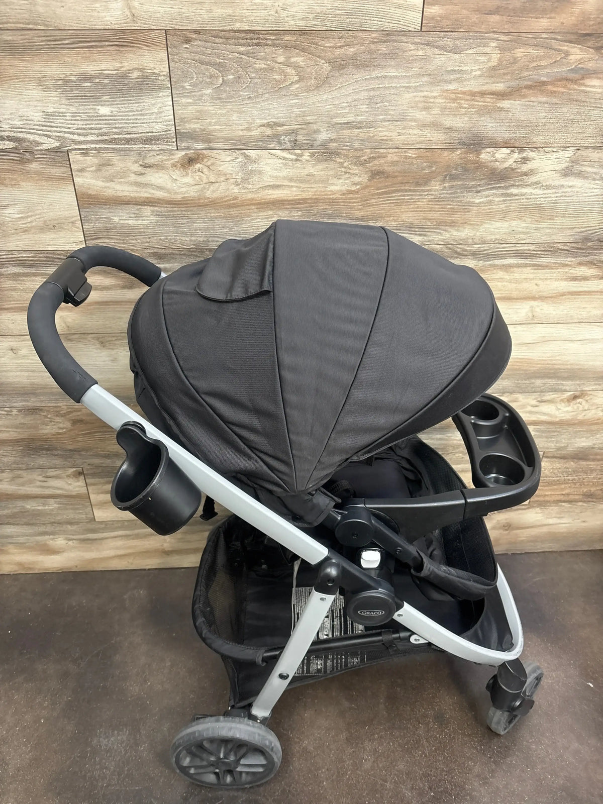 Three-quarter view of Graco Modes Pramette Stroller, Black showing canopy, cup holders, and storage basket on wood floor