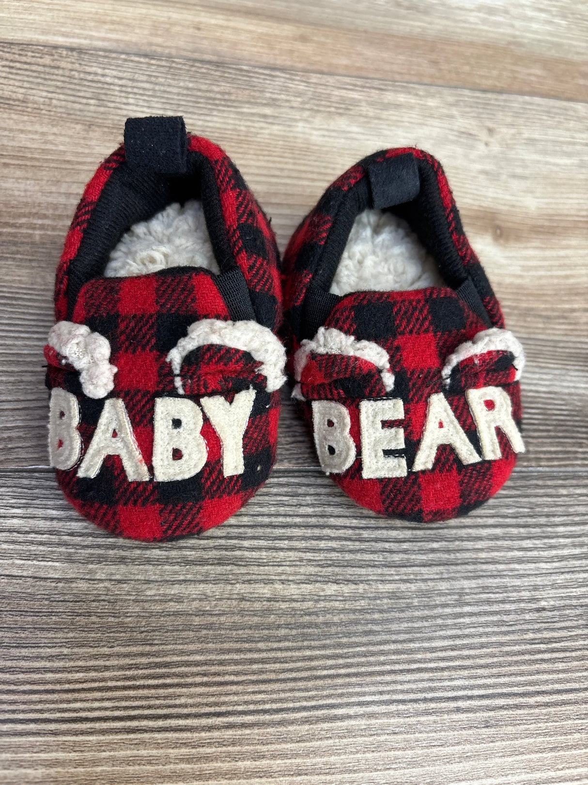 Red plaid baby slippers with fuzzy lining and 'Baby Bear' text on wood background