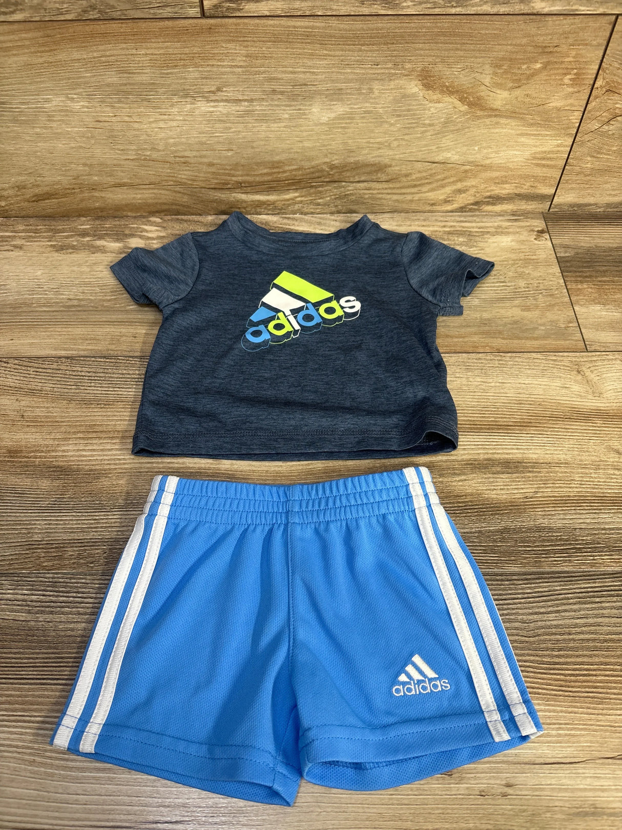 Flat lay of boys' Adidas two-piece set showing navy logo tee and light blue shorts with white side stripes on wood background