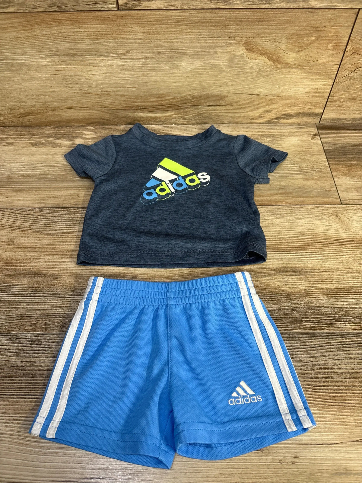 Flat lay of boys' Adidas two-piece set showing navy logo tee and light blue shorts with white side stripes on wood background