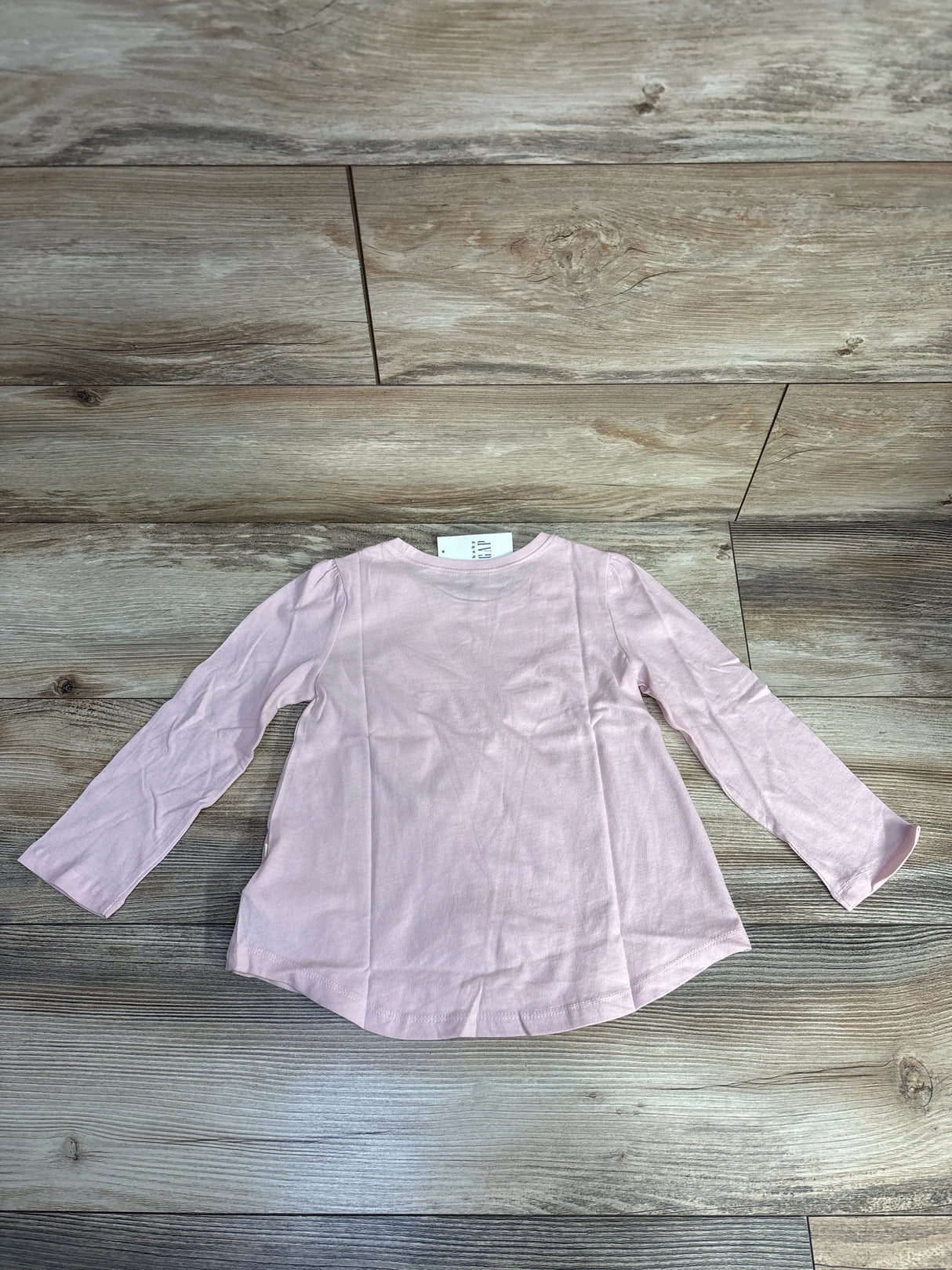 Light pink long-sleeve toddler shirt laid flat on wooden floor, children's clothing