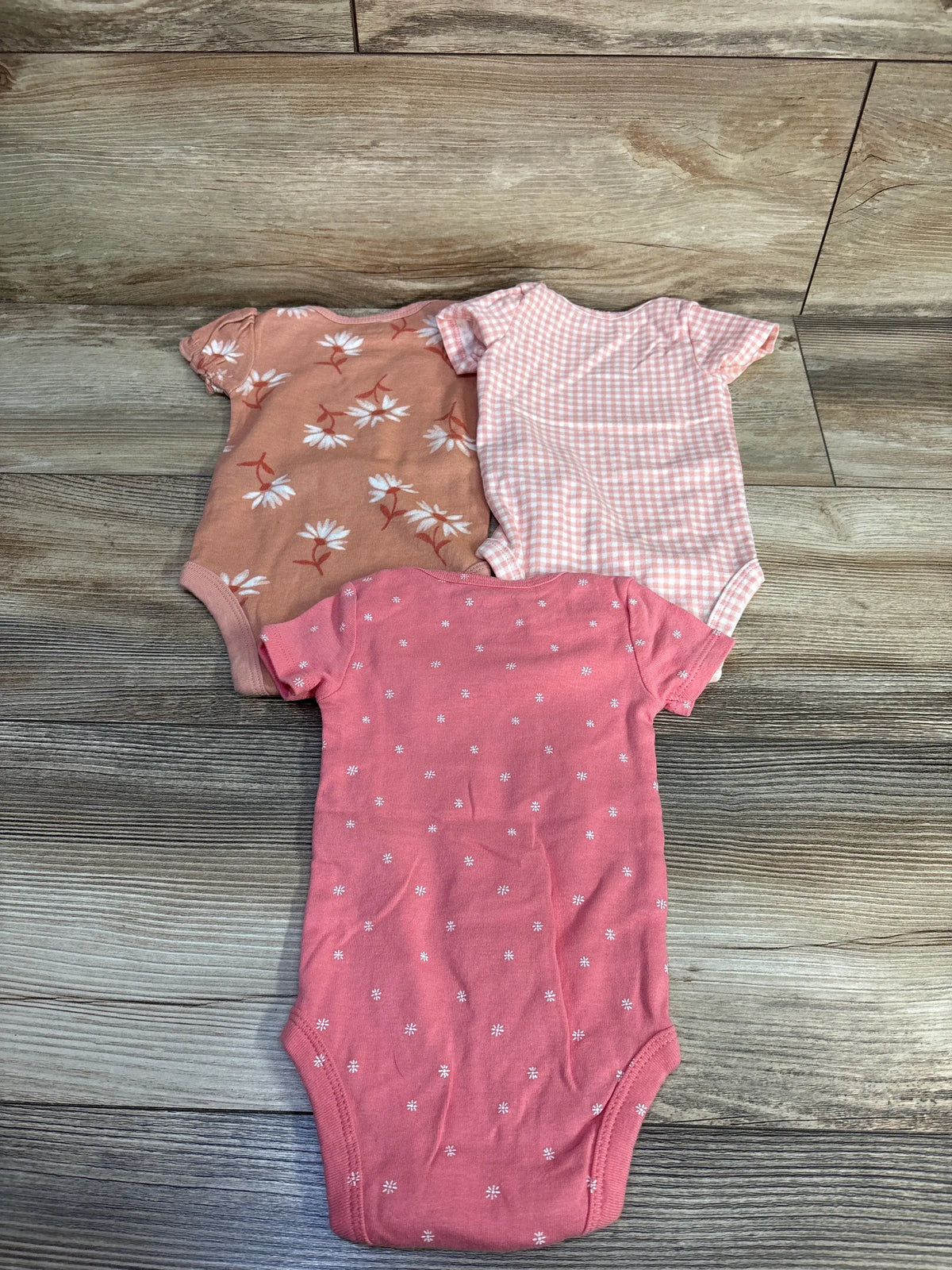 Top view of Just One You three-piece pink baby bodysuit set in size 3 months, showing floral, gingham, and daisy patterns on a wood-look floor