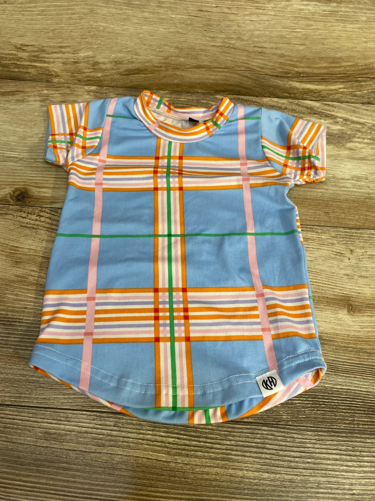 Front view of toddler blue plaid short-sleeve shirt with multicolor stripes, curved hem and brand tag on wood background