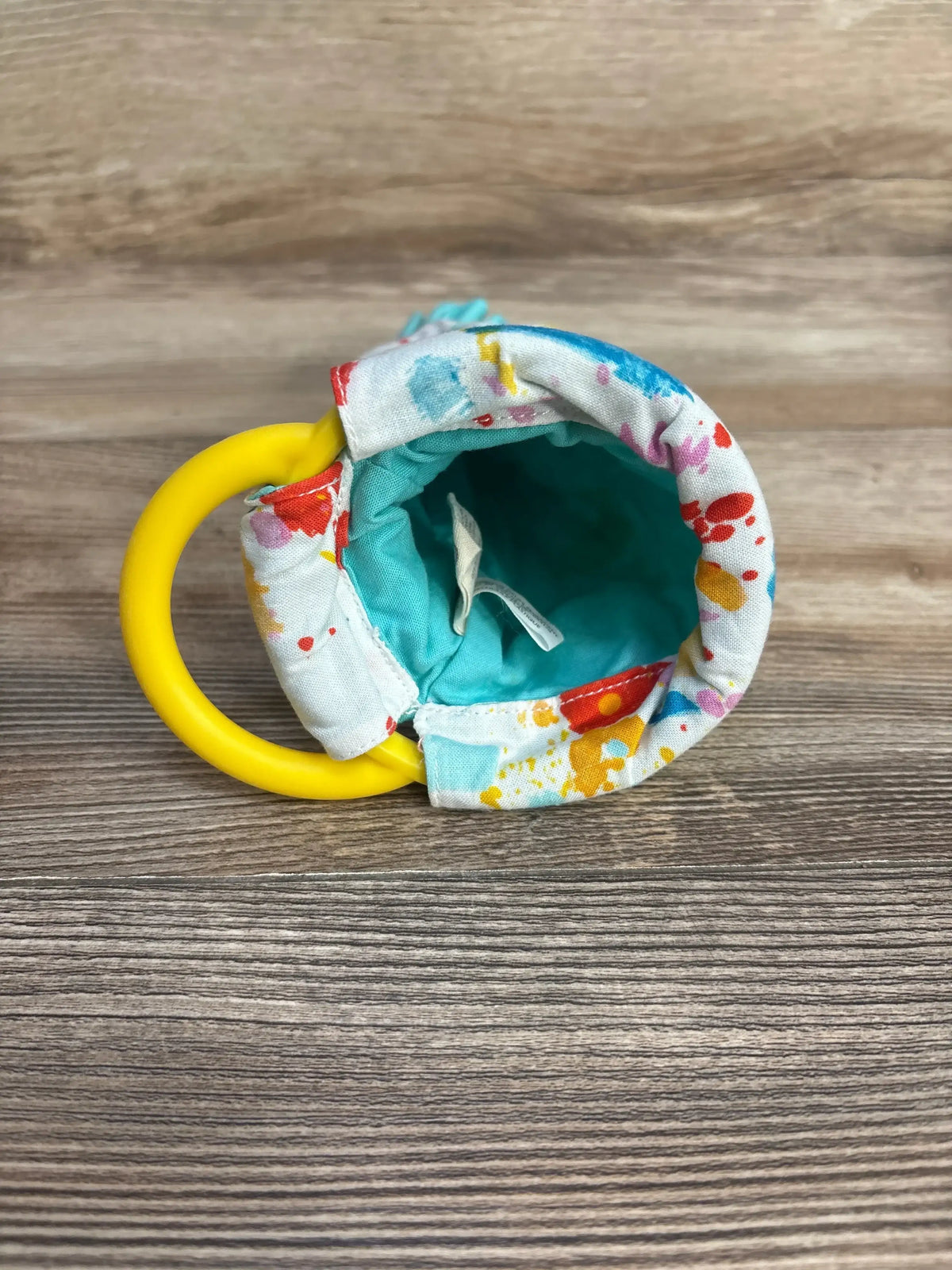 Top view of Lovevery sensory pouch showing open turquoise interior, paint-splatter and yellow ring handle on wood