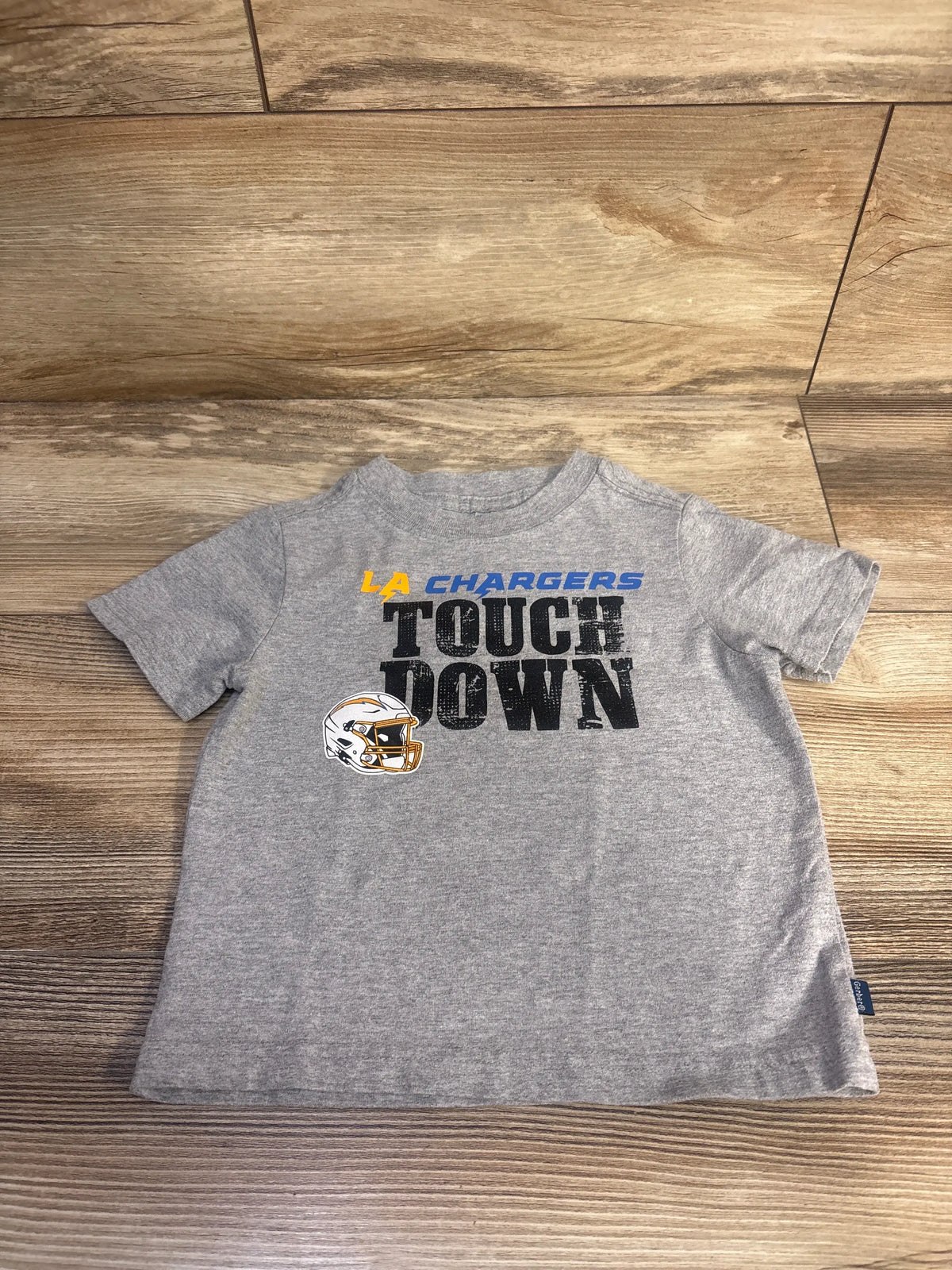 Front view of NFL Team Apparel LA Chargers Touch Down Shirt in grey, showcasing the team graphic and football helmet on wood floor