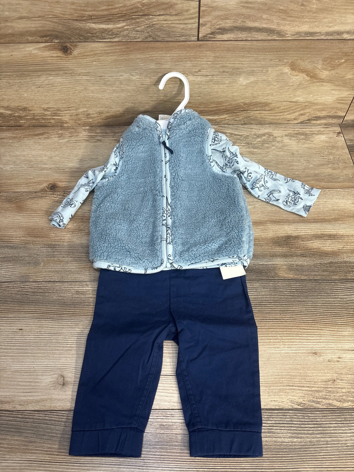 Baby outfit with blue dinosaur print long sleeve shirt, light blue sherpa vest, and navy pants on wooden floor