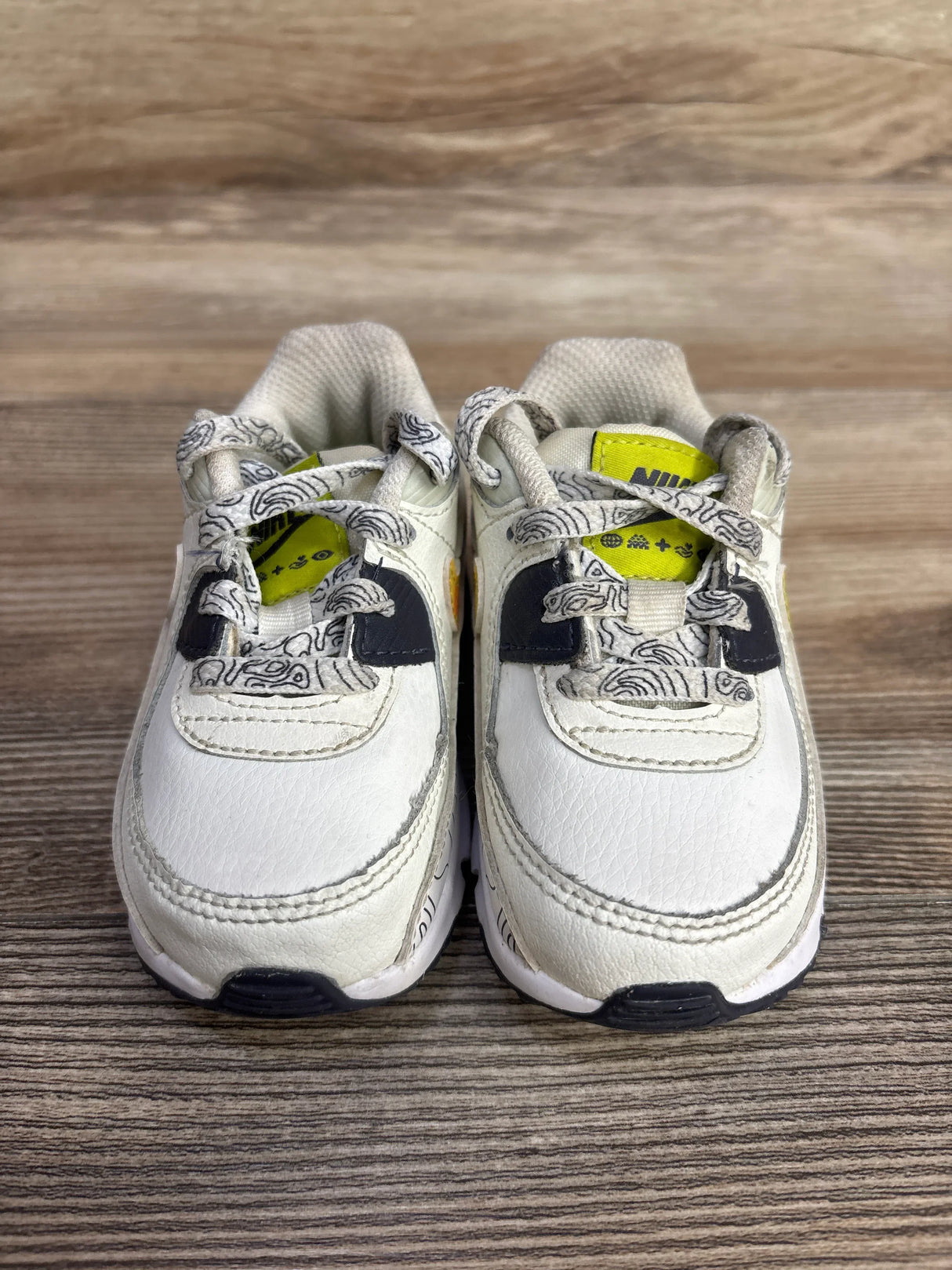 Pair of white Nike toddler sneakers with patterned laces and yellow logo tags on wooden floor