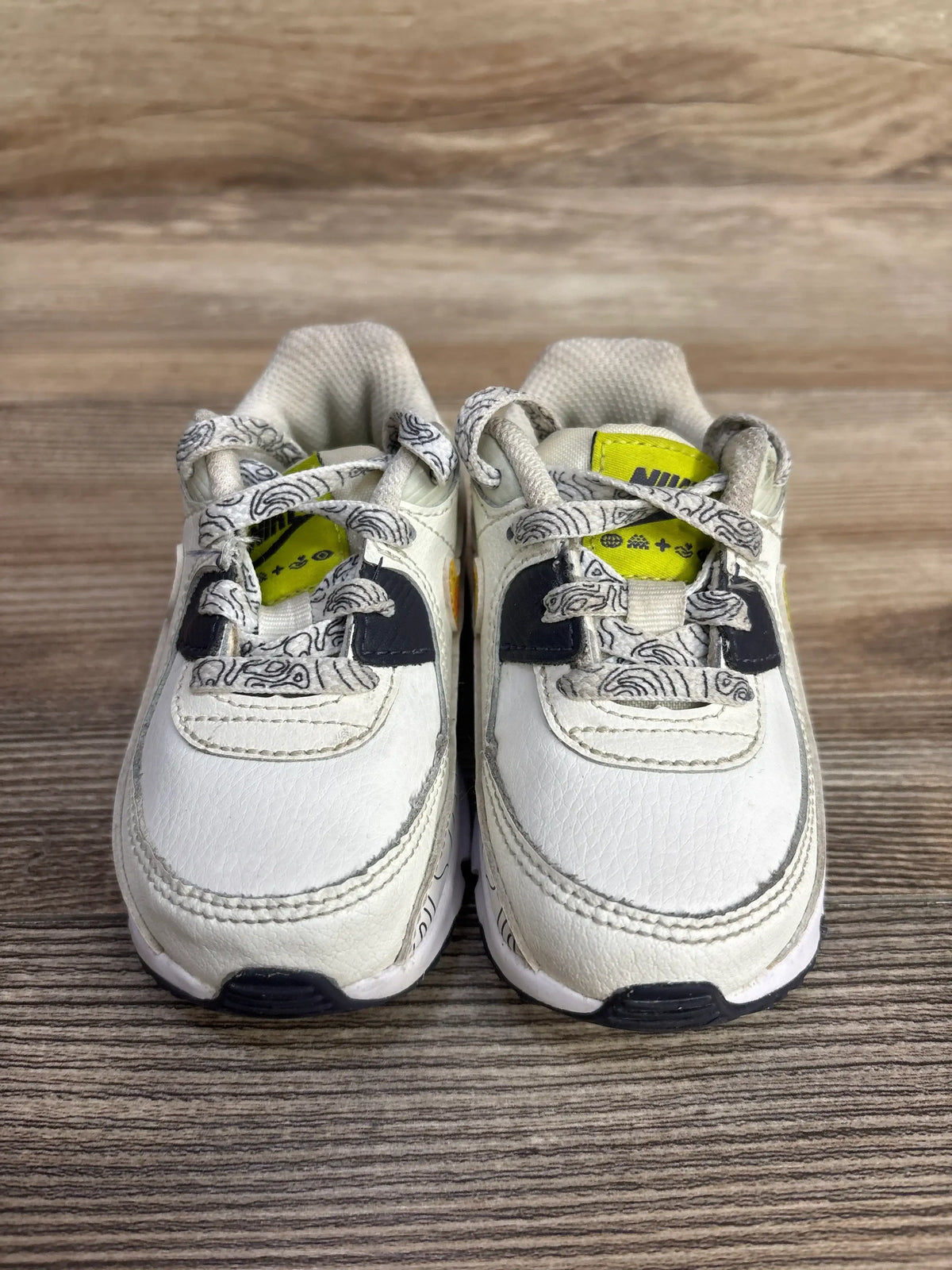 Front view of Nike Air Max 90 toddler sneakers size 4.5C with white leather upper, patterned laces, and yellow Nike tongue tags on wooden floor