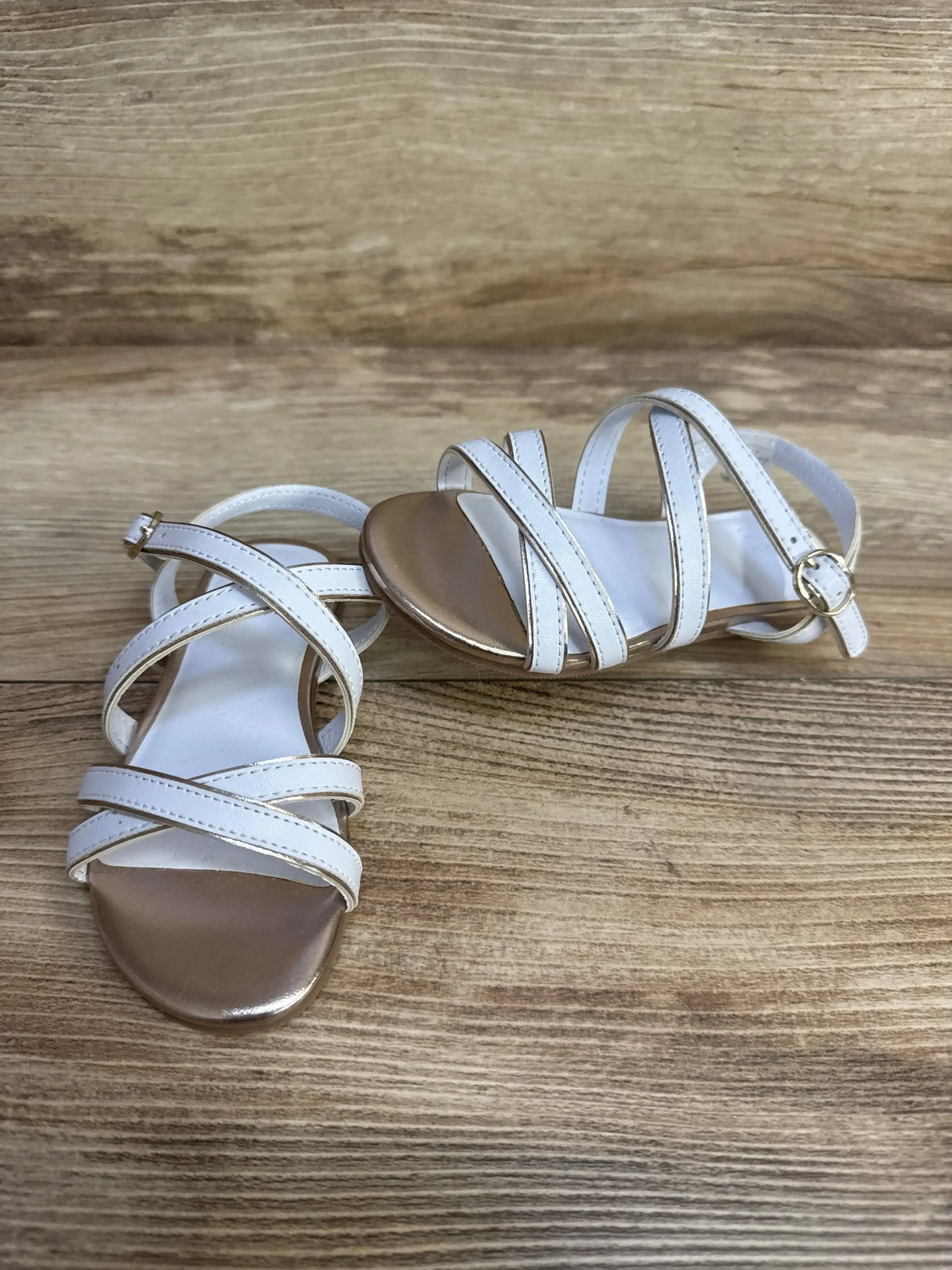 Three quarter view of Janie and Jack white metallic trim sandal with crisscross straps and gold toe on wood background