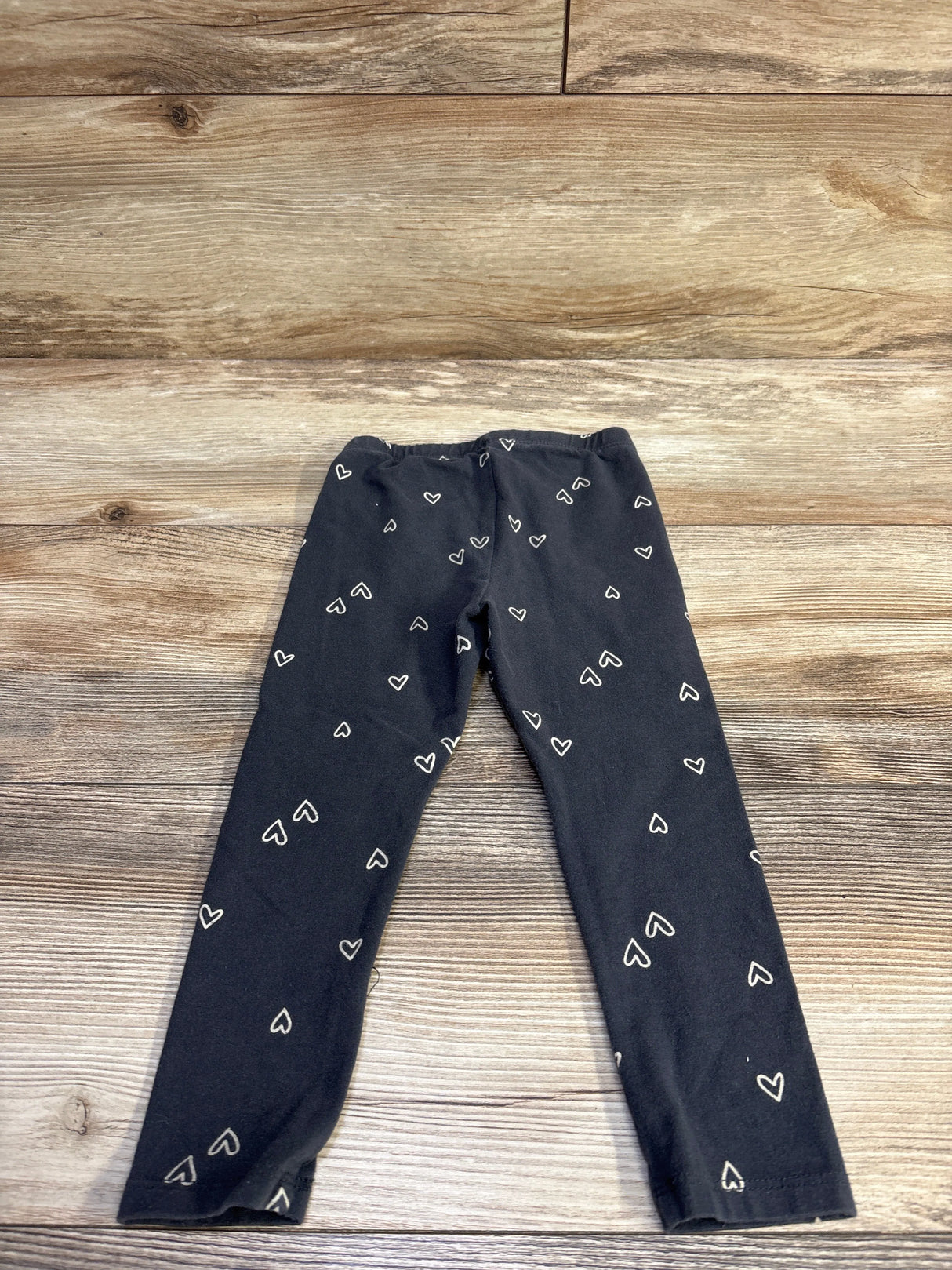 Top-down view of Cat and Jack toddler leggings size 2T with white heart outlines on grey fabric against wood background