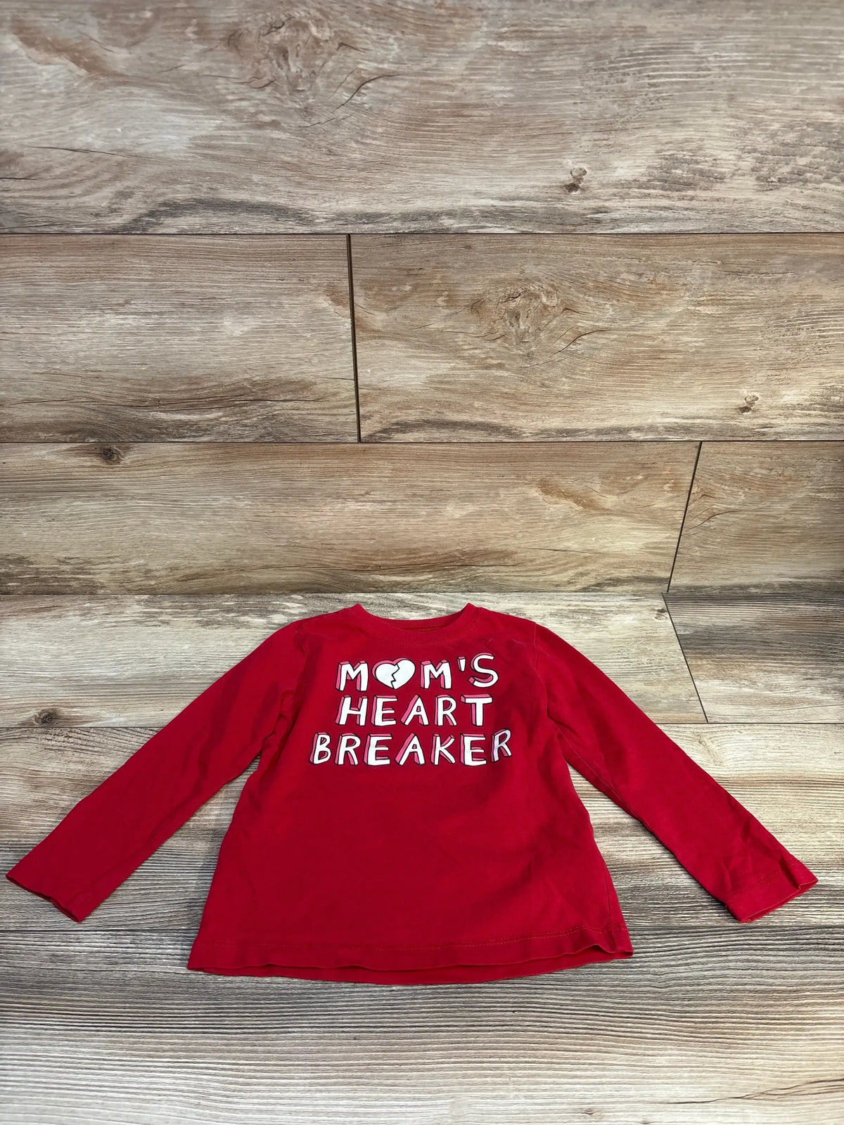 Red long sleeve toddler shirt with 'Mom's Heart Breaker' text, laid on wood floor