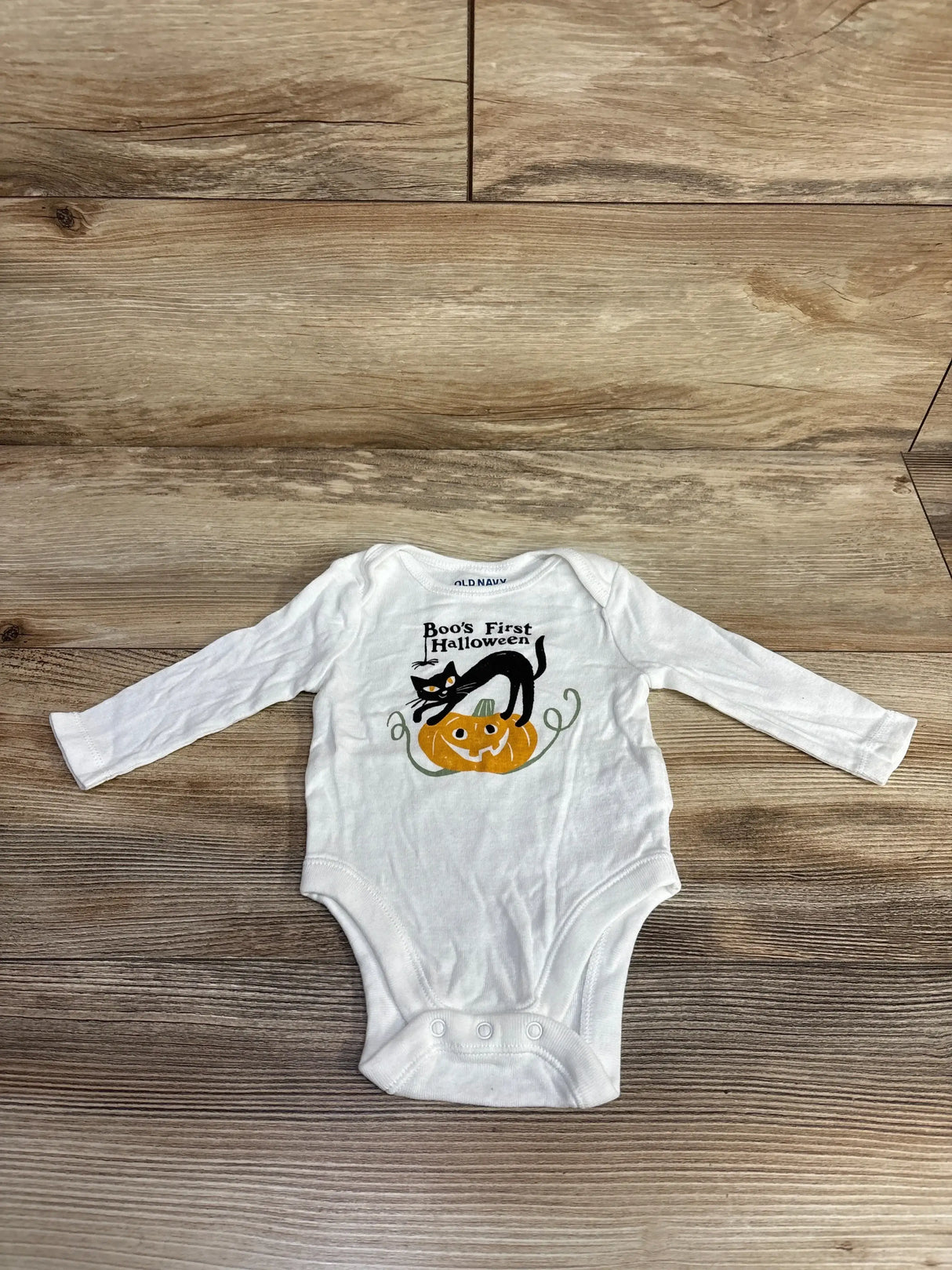 White baby onesie with 'Boo's First Halloween' design, black cat, and pumpkin on wood floor