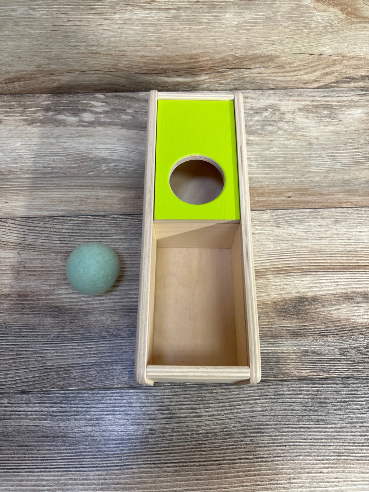 Wooden toy box with green top featuring circular hole and a blue felt ball on wooden surface