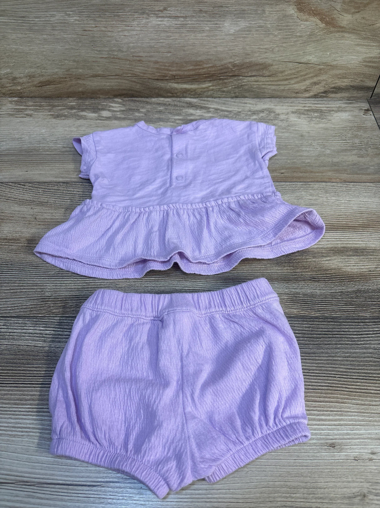 Flat lay of purple Carter’s 2-piece peplum top and elastic-waist shorts showing ruffled hem and back snaps on wood background