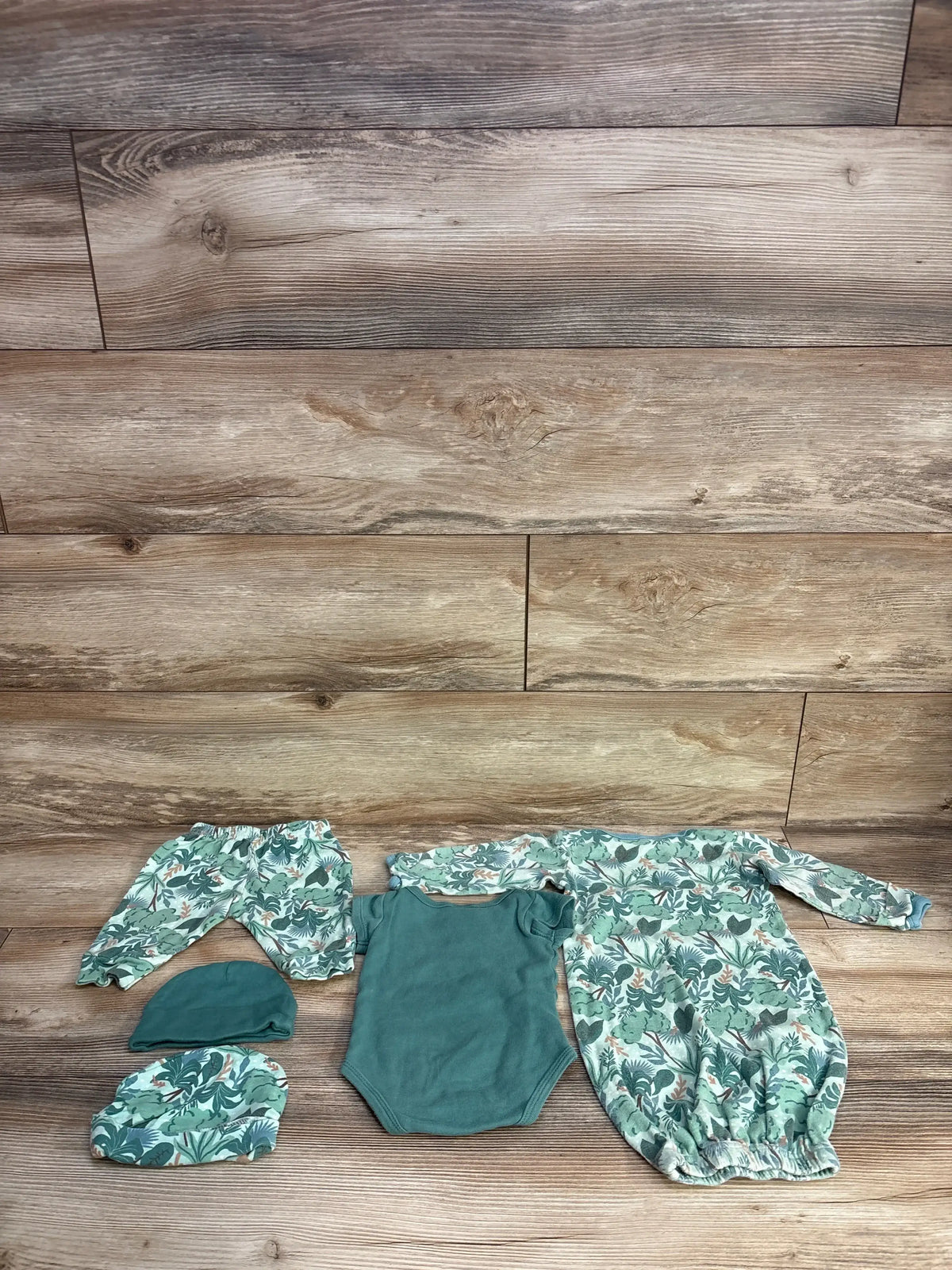 Top-down view of 5-piece newborn Peanutshell set: green onesie, leaf-print gown, pants and two matching hats on wood floor