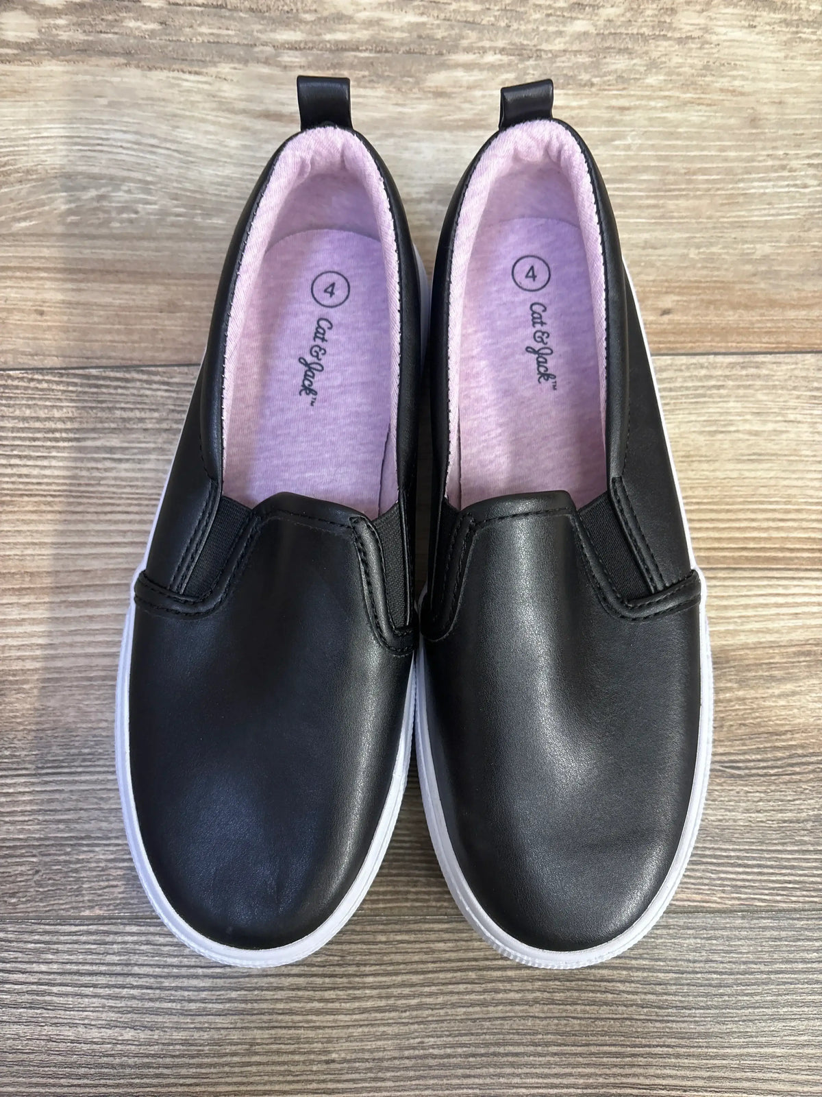 Top-down view of Cat and Jack Hailey slip-on toddler sneakers in black showing white rubber soles and pink fabric insoles on wood floor