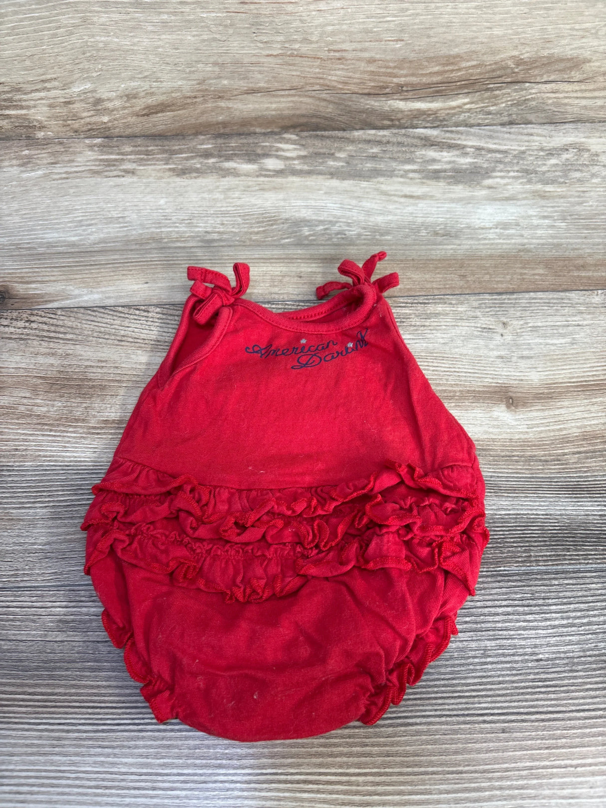 Top-down view of red newborn bubble romper with American Darling script showing shoulder tie straps and layered ruffles on wood background