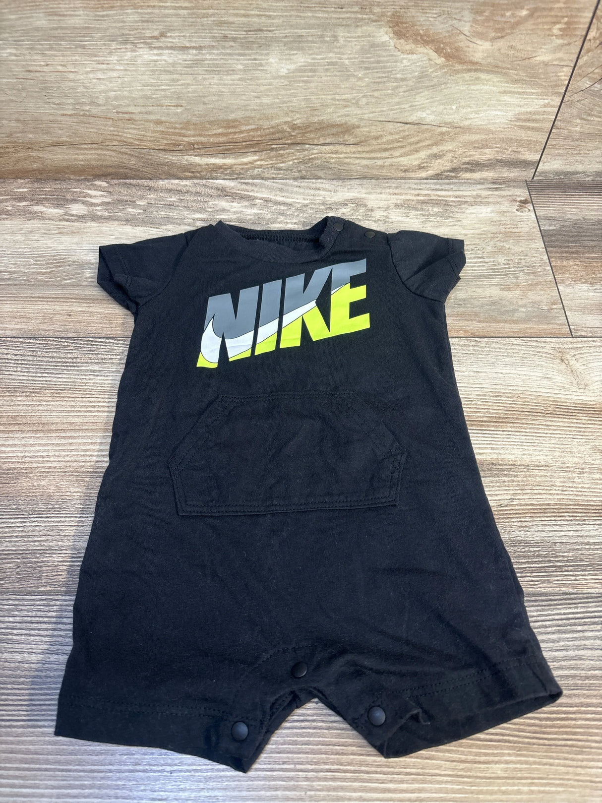 Black short sleeve baby romper with NIKE logo, front pocket, snap bottom, on wood floor