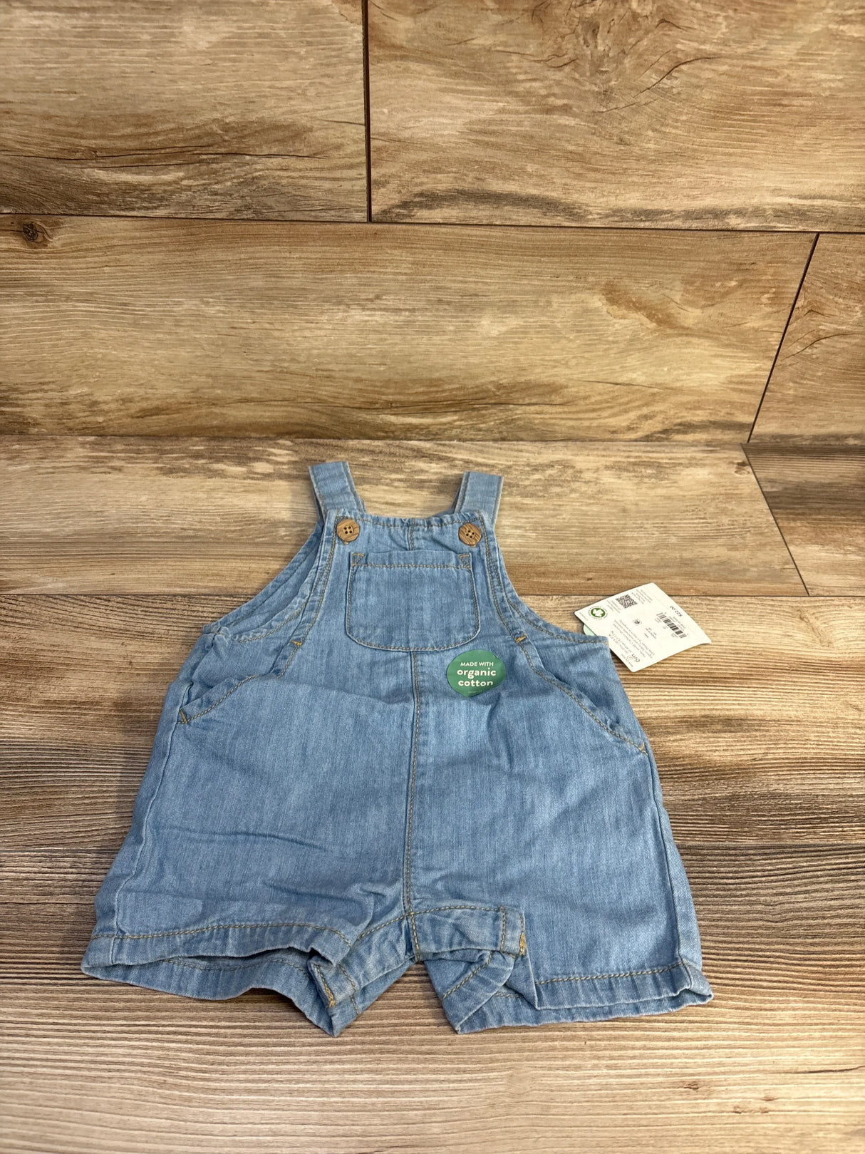 Front view light blue chambray shortall, size 6m, chest pocket, wooden strap buttons, organic cotton tag on wood
