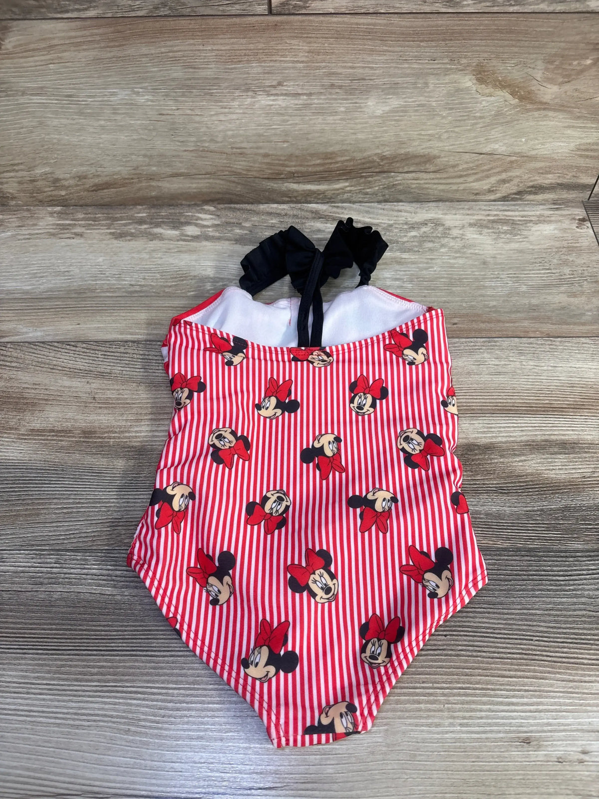 Front of toddler red Minnie Mouse one-piece swimsuit with red and white stripes, Minnie faces, and black ruffle halter straps on wood floor