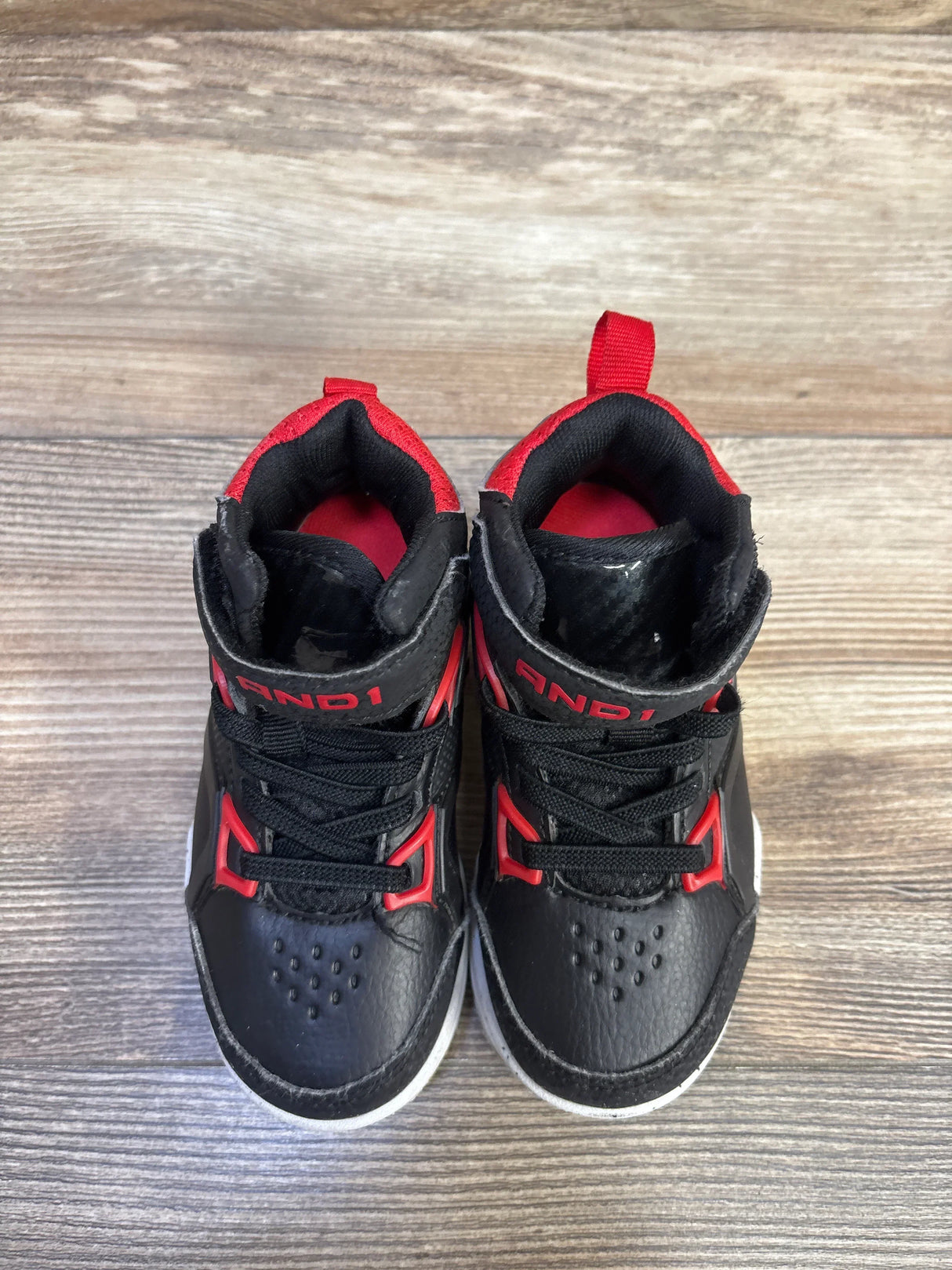 Top-down view of toddler AND1 black basketball sneakers with red accents, lace-up front and Velcro ankle strap on wood floor