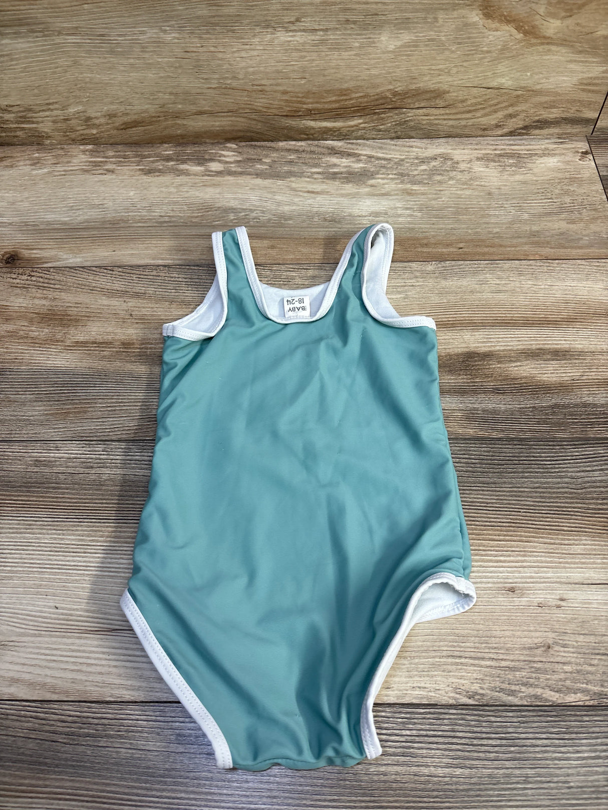 Top-down view of Cotton On baby swimsuit in green showing white trim and size 18-24 months tag on wood background