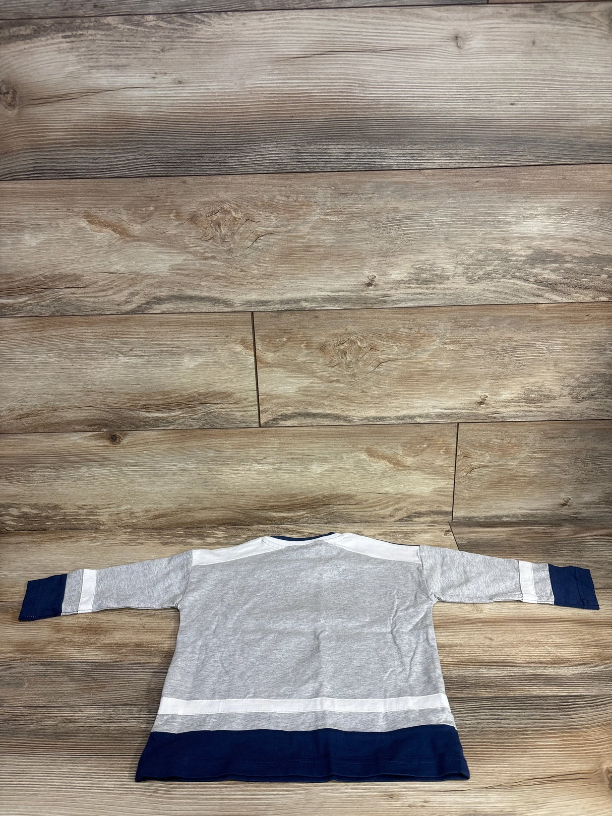 Back view of Cotton On Kids Golden State Warriors toddler long-sleeve top (2T) showcasing gray body with white stripe accents and navy cuffs and hem on wood floor