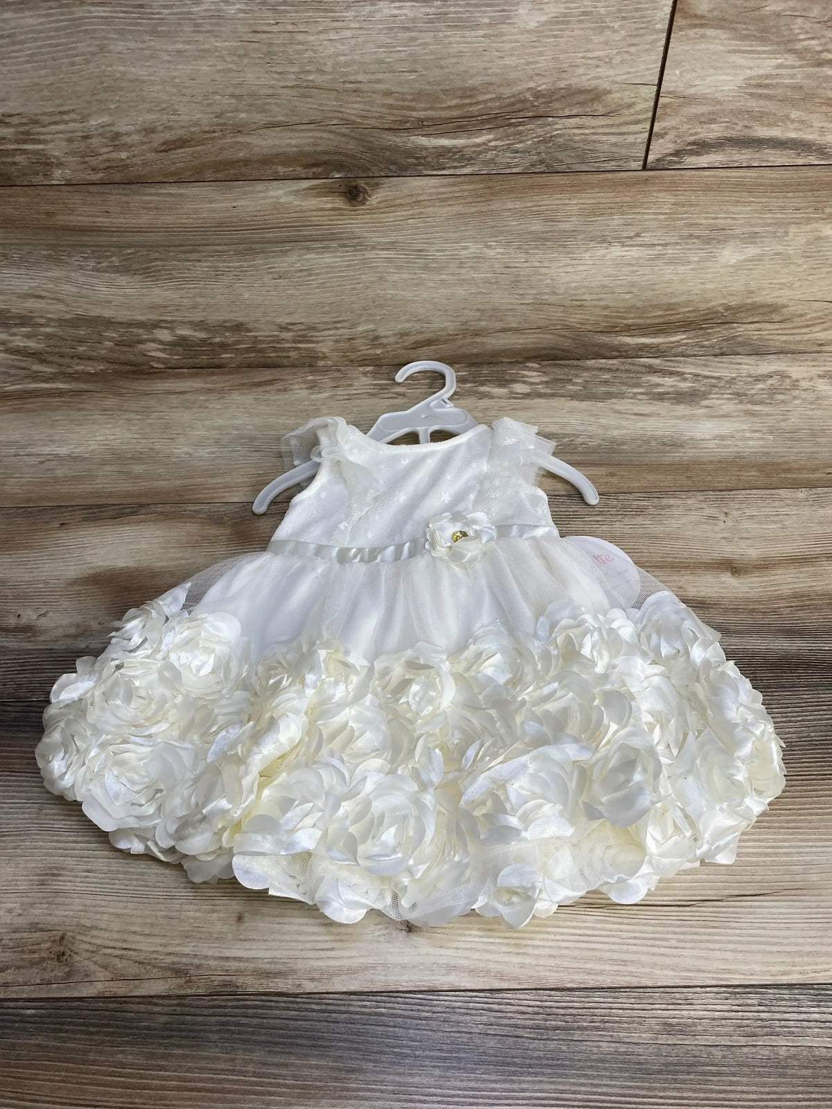 Front view of ivory baby rosette dress with layered rosette skirt, satin waist ribbon, tulle bodice on hanger over wood background