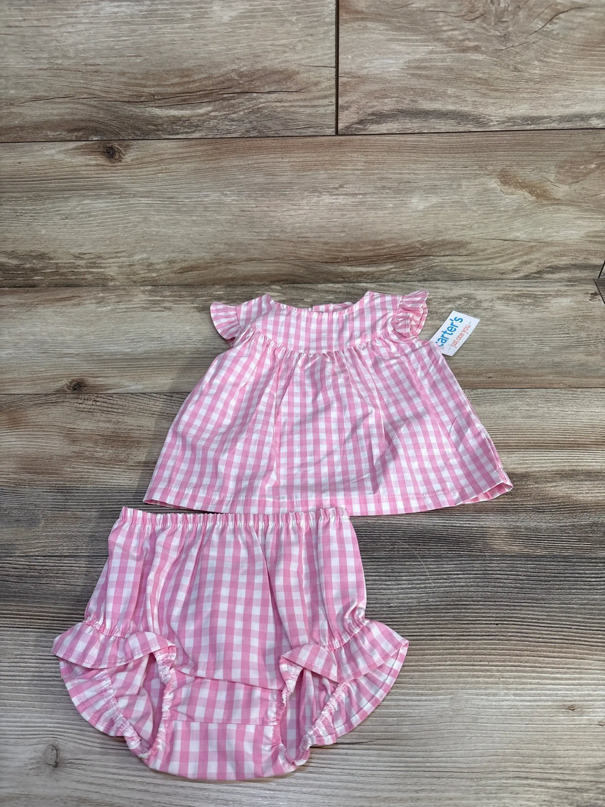 Top-down view of a pink gingham 100% cotton sleeveless top and matching bloomers with ruffled edges on a wood surface