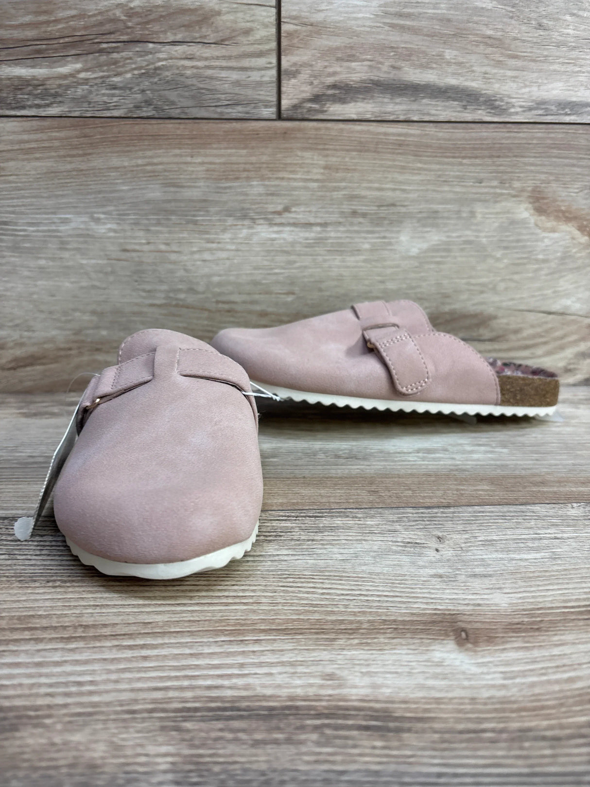Pink suede slip-on clogs with adjustable buckle and white soles on wooden floor