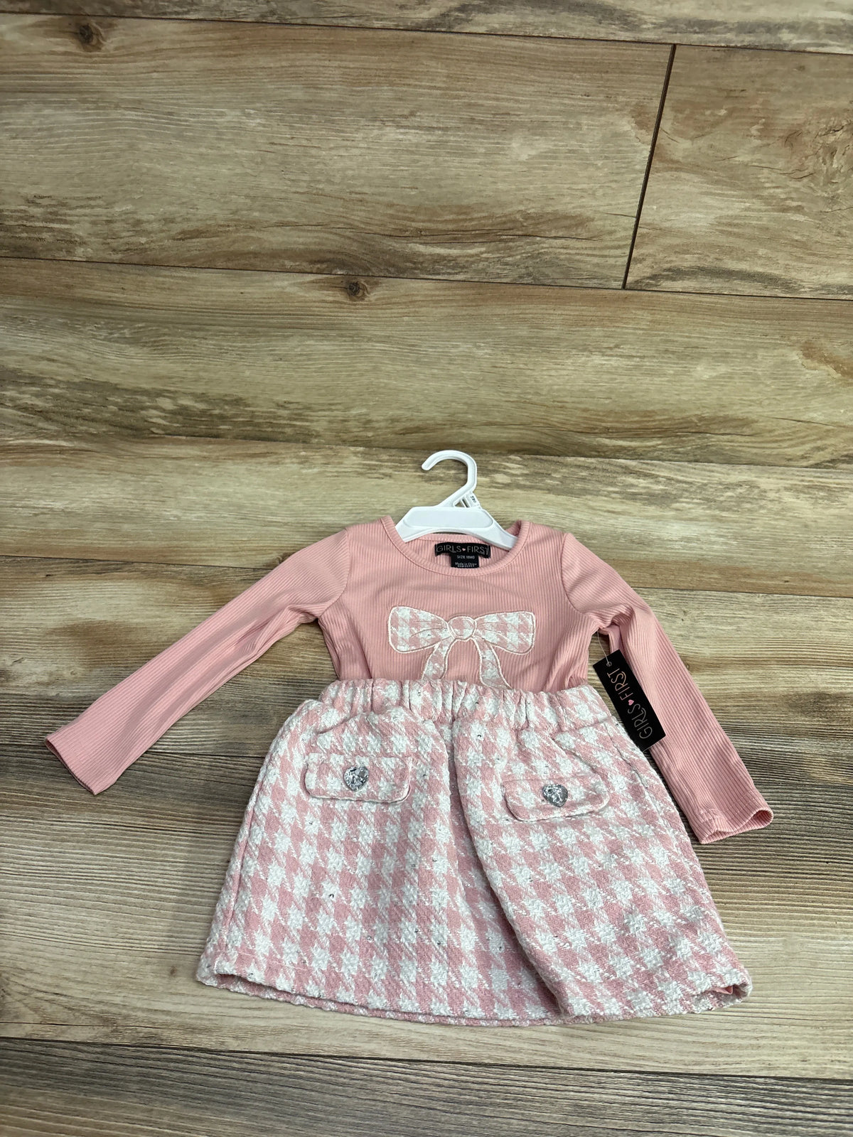 Front view of pink Girls First long sleeve dress with ribbed sleeves, glittery bow, and patterned skirt on a hanger
