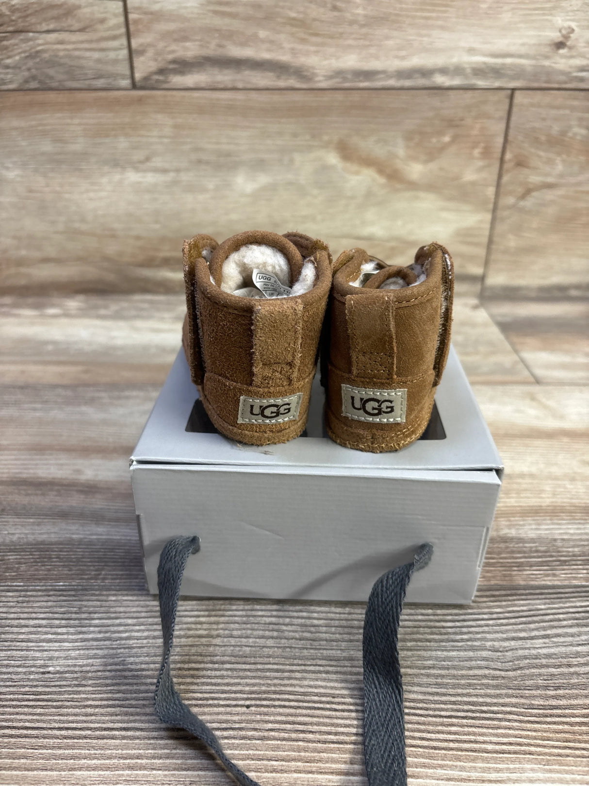 Rear view of UGG Baby Neumel booties showing heel UGG logo and plush shearling lining on a white box with wood backdrop