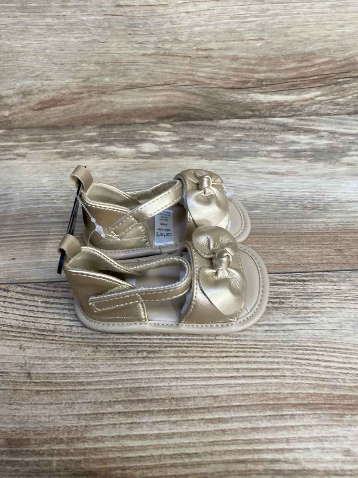 Top-down view of Child of Mine gold baby sandals showcasing knotted bow straps and hook-and-loop ankle closure on wood background