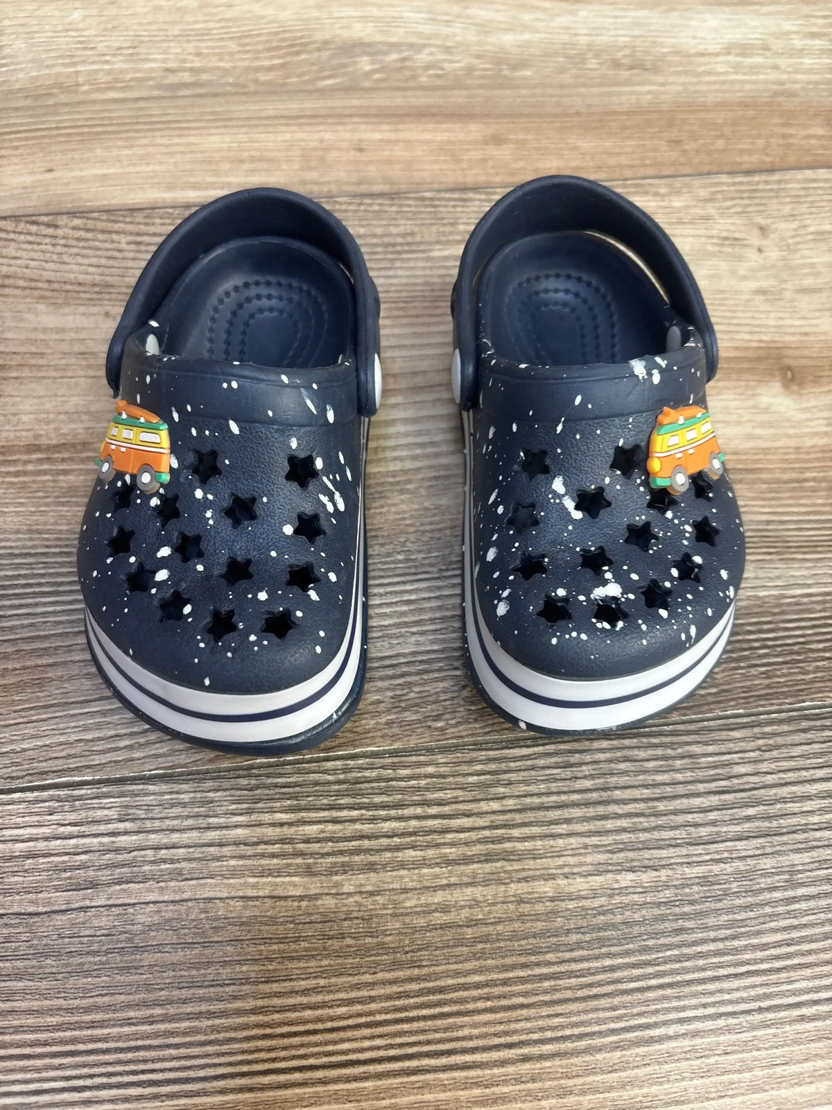 Top-down view of navy toddler clogs (size 4C) showcasing star-shaped cutouts, white paint-splatter pattern, striped white soles, and orange camper charms on a wood floor