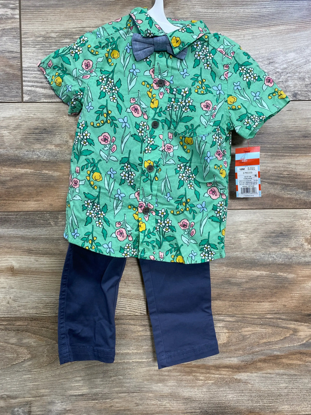 Front flat lay of Cat and Jack 3-piece floral set showing green button-up shirt with bowtie and navy pants on wood background
