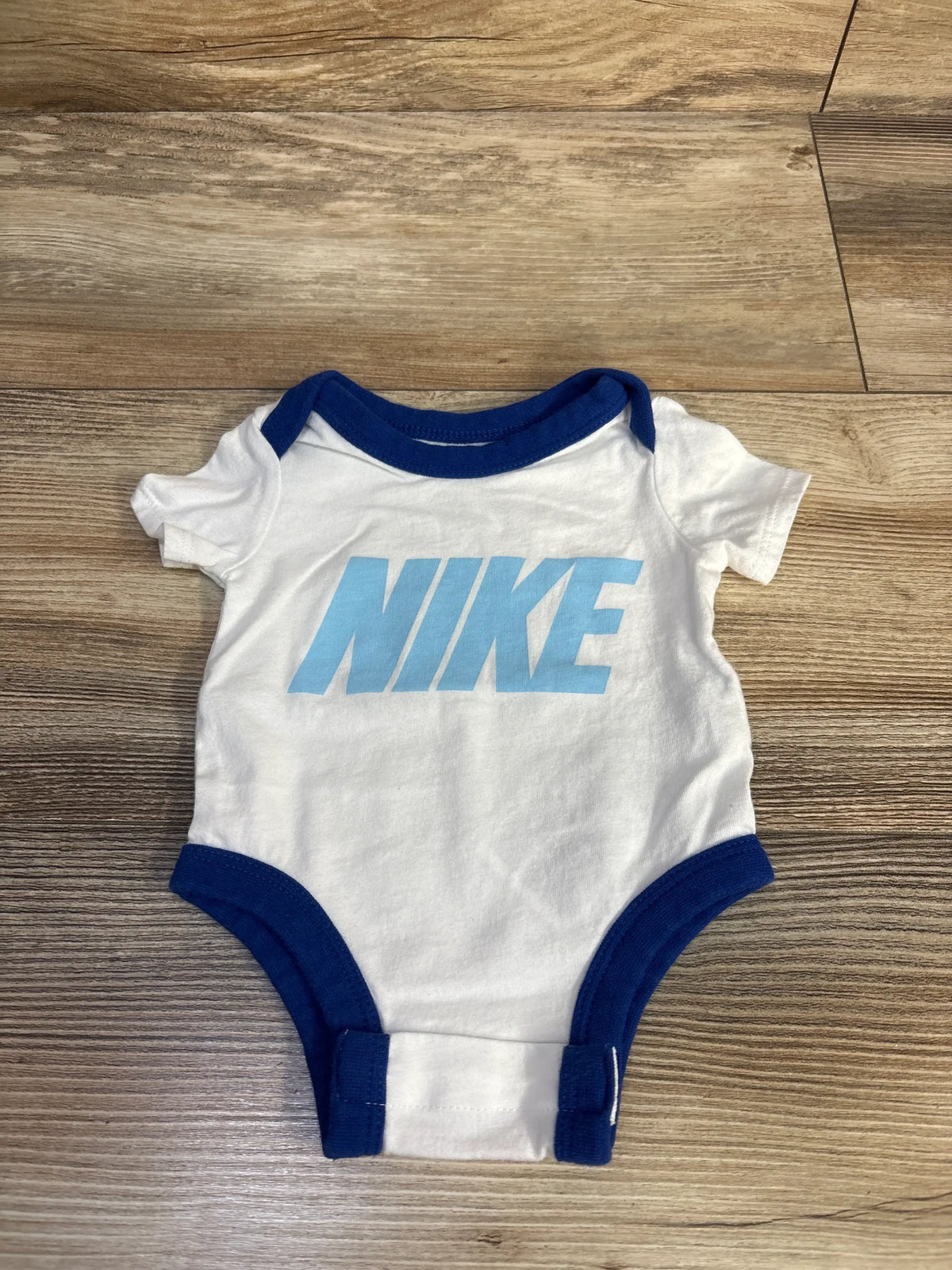 Flat lay of newborn white Nike bodysuit showing light blue NIKE logo, navy trim, and snap crotch on wood floor