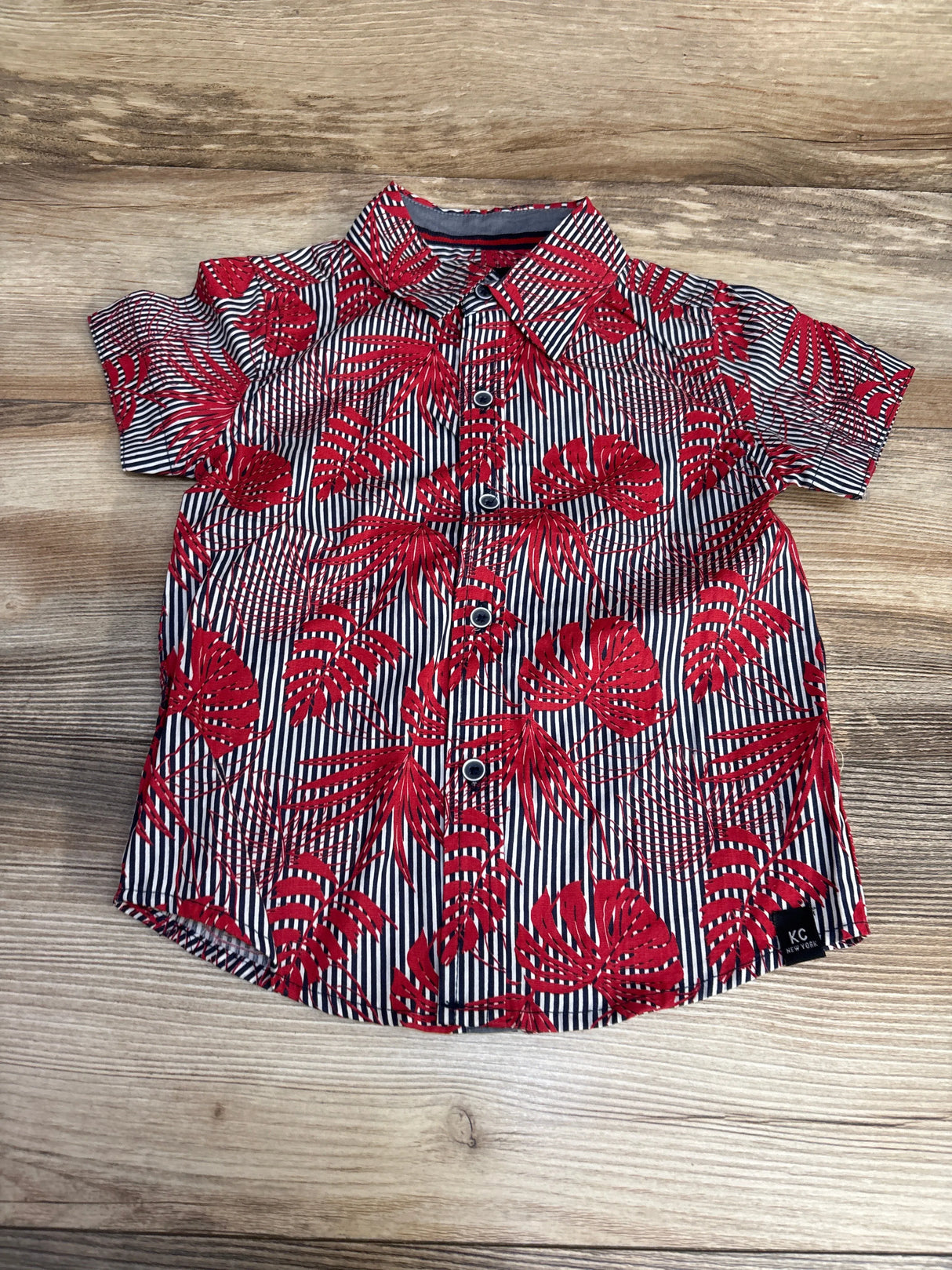 Red tropical leaf print short sleeve button-up shirt for kids on wooden surface