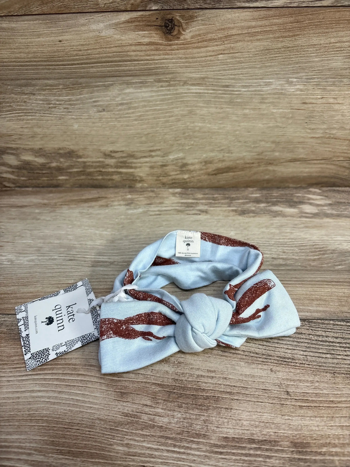 Top view of Kate Quinn bow headband showcasing light blue fabric with orange fox print and brand hangtag on wood plank floor