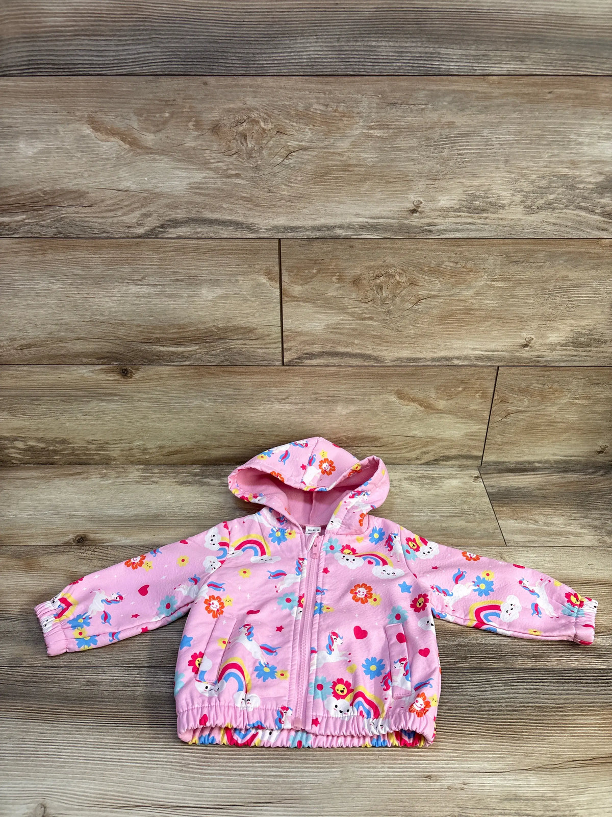 Front view of pink hooded baby jacket with unicorn and rainbow print, zipper and elastic hem on wood background