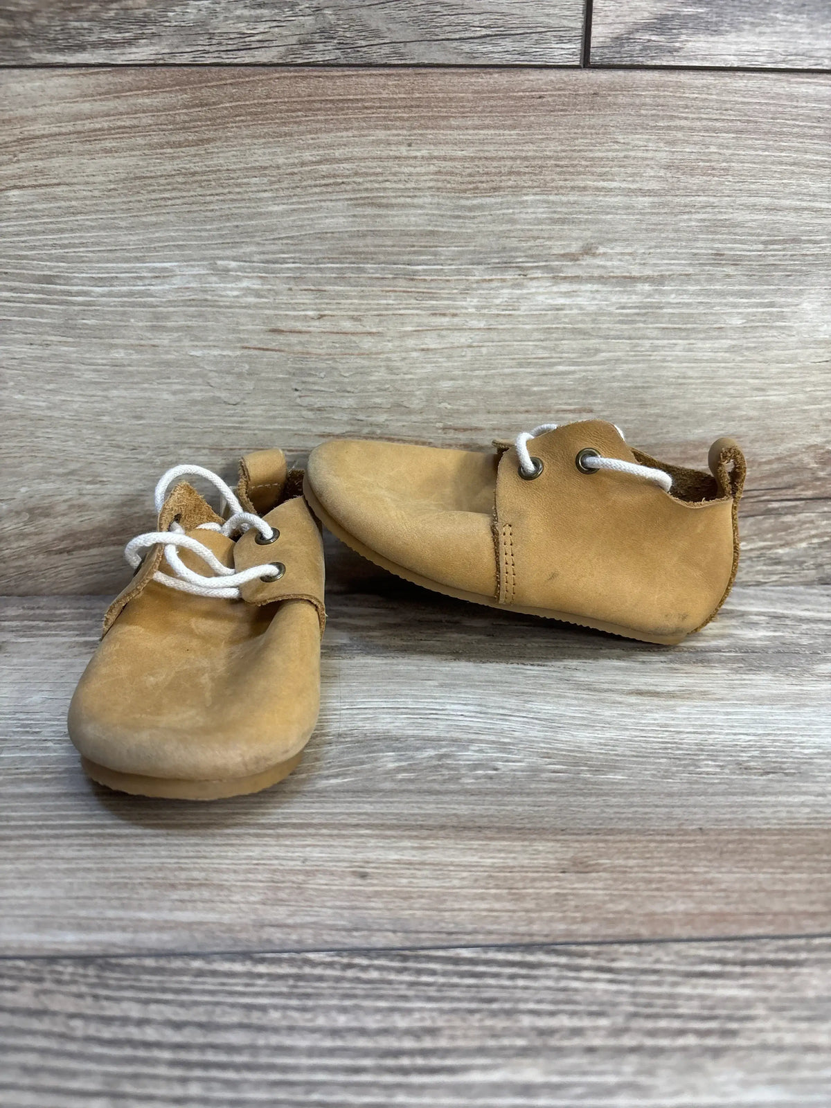 Angled view of Piper Finn Natural low-top oxford baby shoes, size 6C, tan suede with white laces, gently used, on wooden background