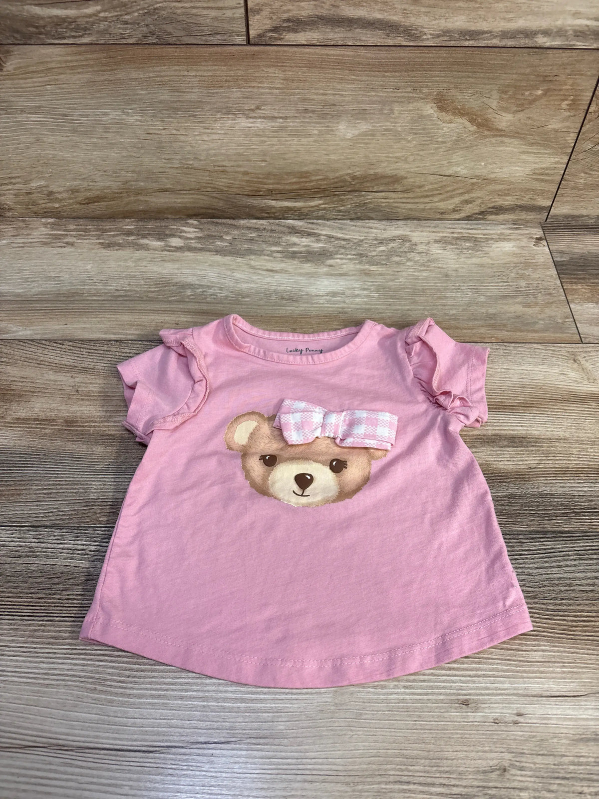 Front view of pink Lucky Penny bear shirt, size 2T, with ruffled sleeves and bear graphic on wood-look floor