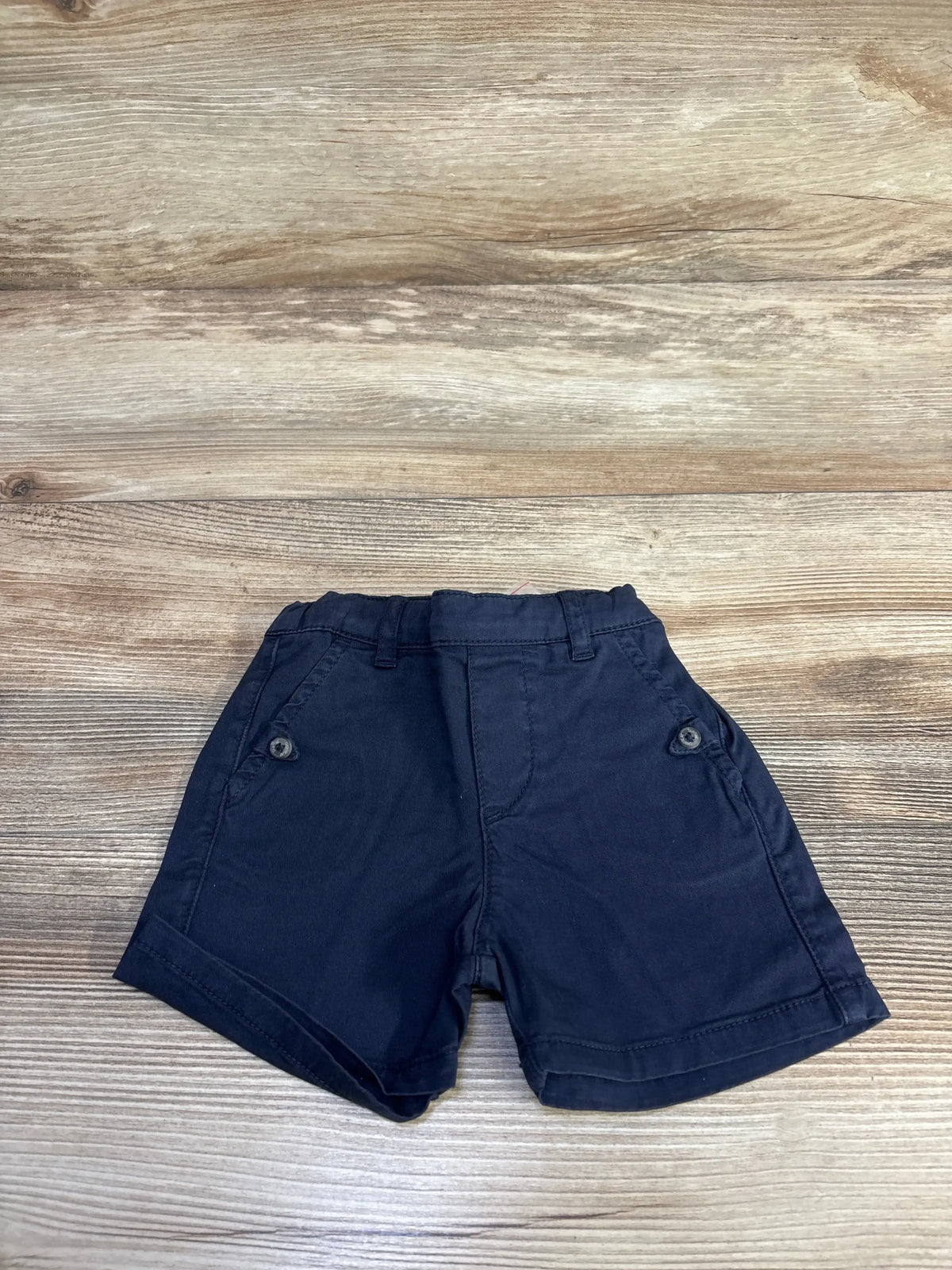 Flat lay front view of navy toddler chino shorts (12 months) showcasing button-trim side pockets, cuffed hems, and elastic waistband on light wood background