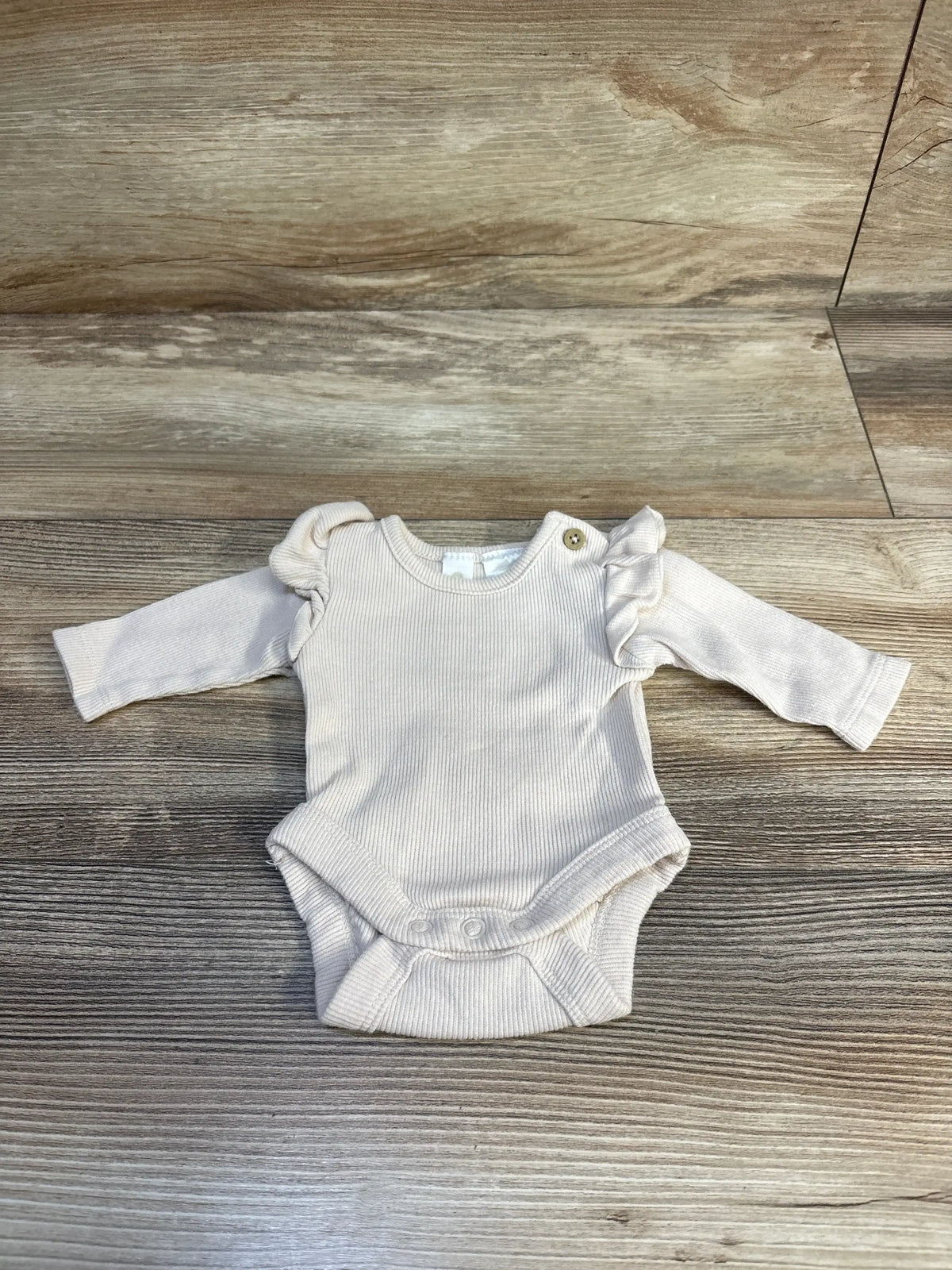 Front view of beige ribbed preemie bodysuit showcasing ruffled shoulder detail and snap crotch on light wood background