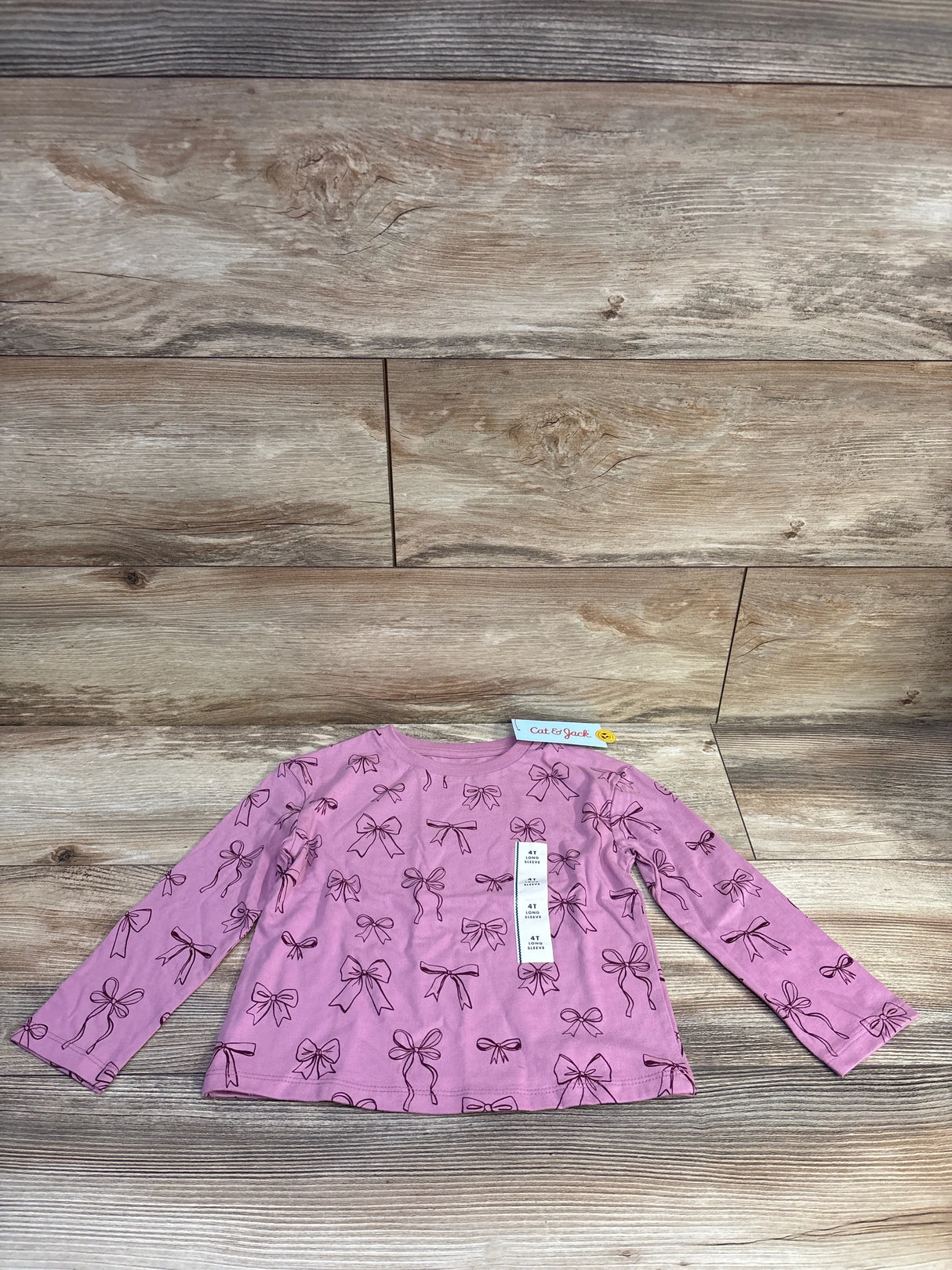 Flat lay of pink Cat & Jack long-sleeve toddler shirt showing black bow print and attached 4T size tag on wood background