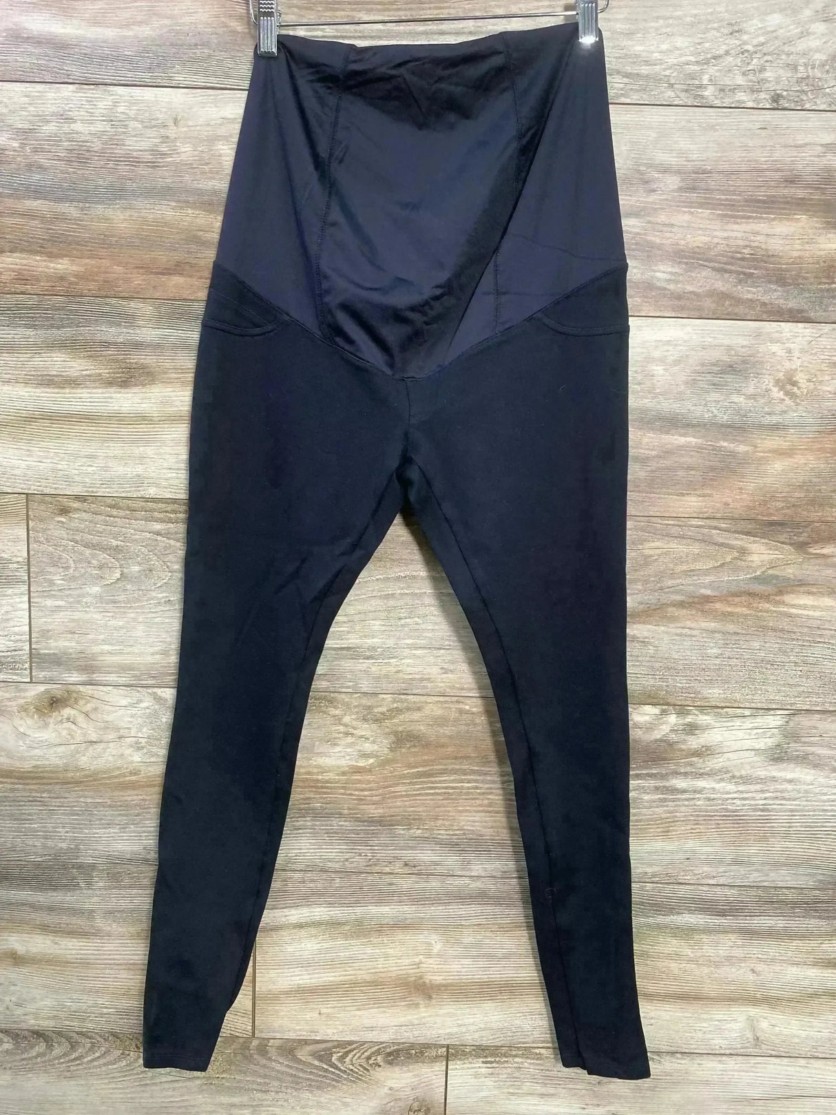 Front view of Isabel maternity crossover panel pants in black showing crossover belly panel and slim tapered legs on wood floor background