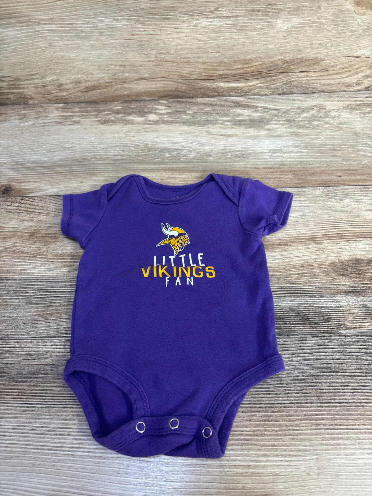 Front view of purple NFL Vikings baby bodysuit showcasing Little Vikings Fan graphic and snap closures on a wood background